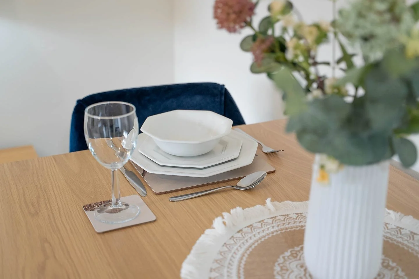 Empty dining table set with a wine glass, stacked white plates, a bowl, a fork, and a spoon, with a vase of flowers in the foreground.