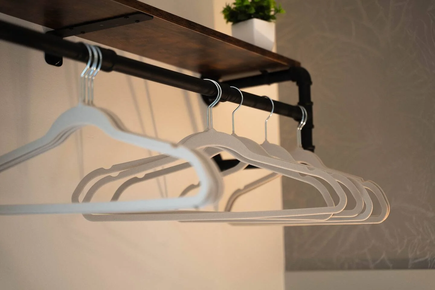A black metal clothing rack with four empty white hangers, situated beneath a wooden shelf with a potted green plant, against a wall with a gray textured section.