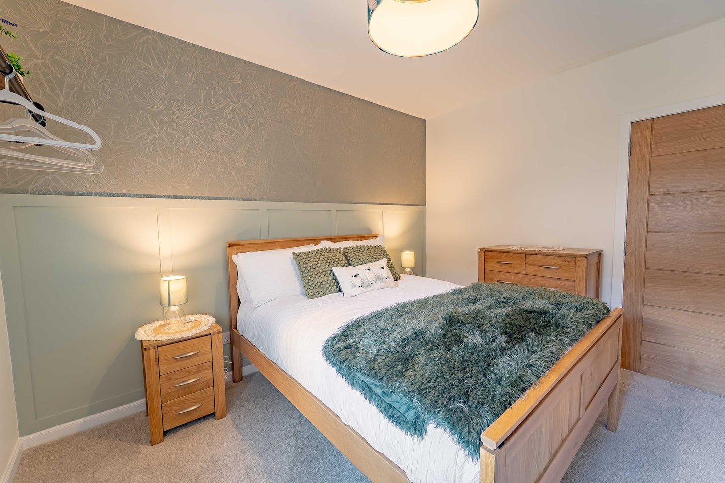 A cozy bedroom with a wooden bed frame, white bedding, green pillows, a plush green blanket, two bedside lamps, a wooden dresser, and a closet door.