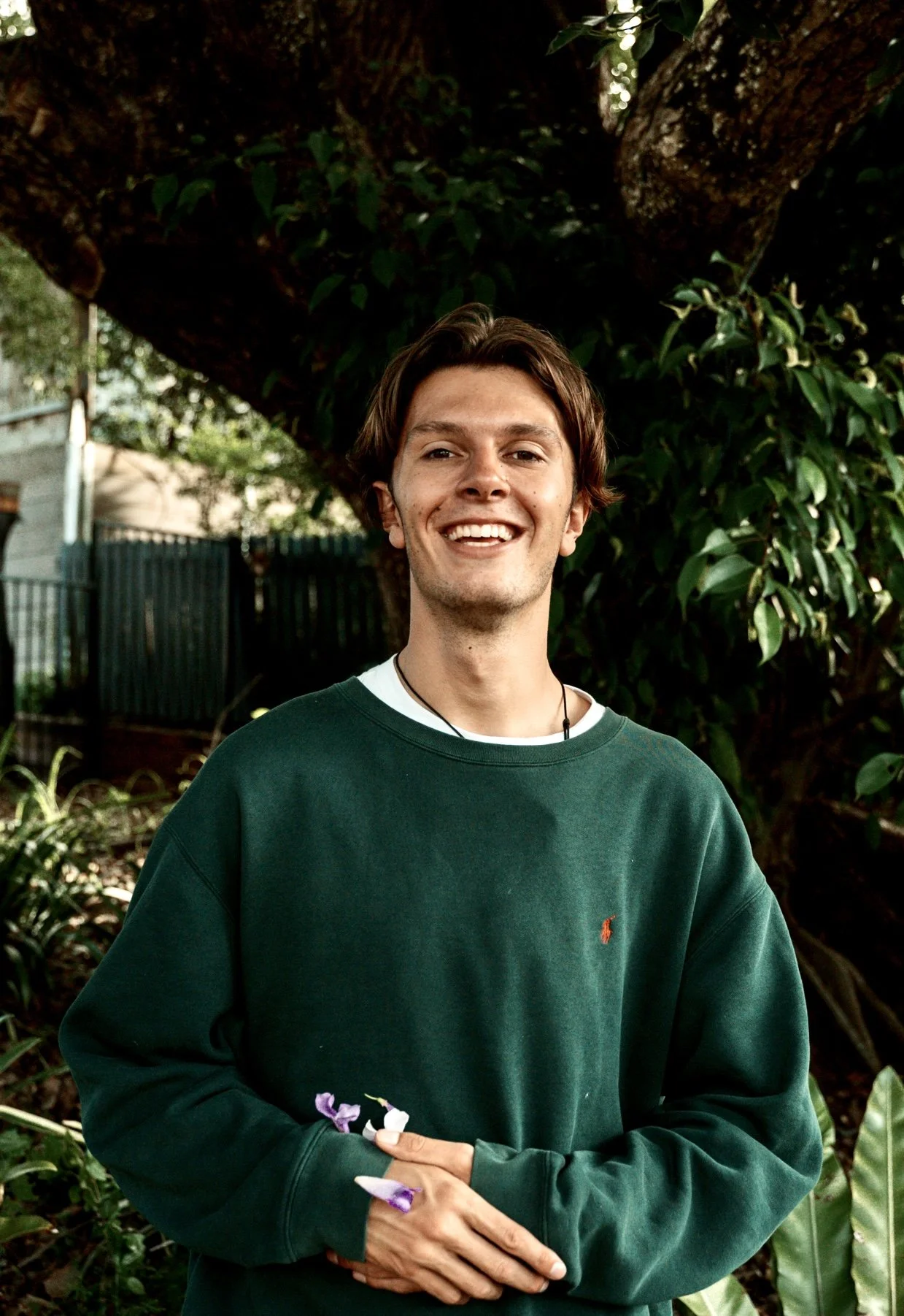 Smiling young man with medium-length brown hair standing outdoors in front of a large tree with dark green leaves. He is wearing a dark green sweatshirt over a white shirt and holding purple flowers in his hand.