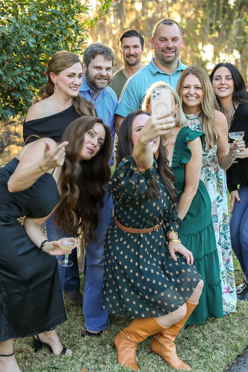 Group of nine friends taking a selfie together outdoors during sunset, some holding drinks, smiling and posing cheerfully.