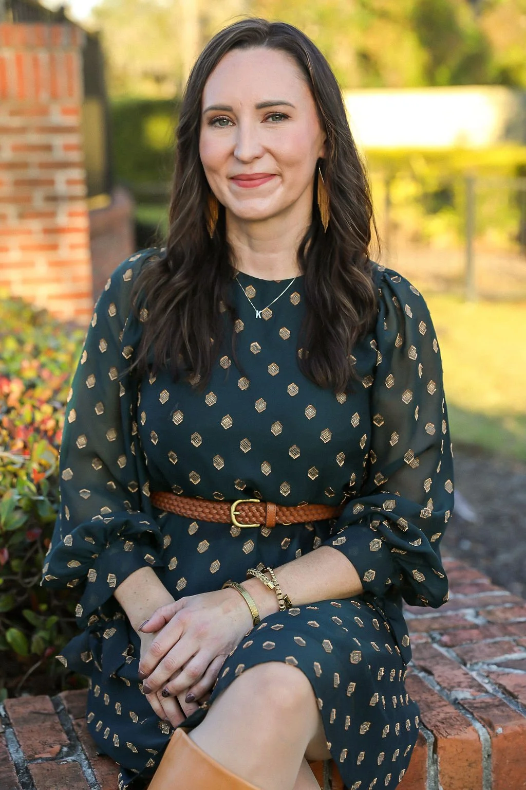 A woman sitting outdoors on a brick ledge, wearing a dark blue dress with gold geometric patterns, accessorized with gold jewelry, and a brown belt, with trees and a brick building in the background.