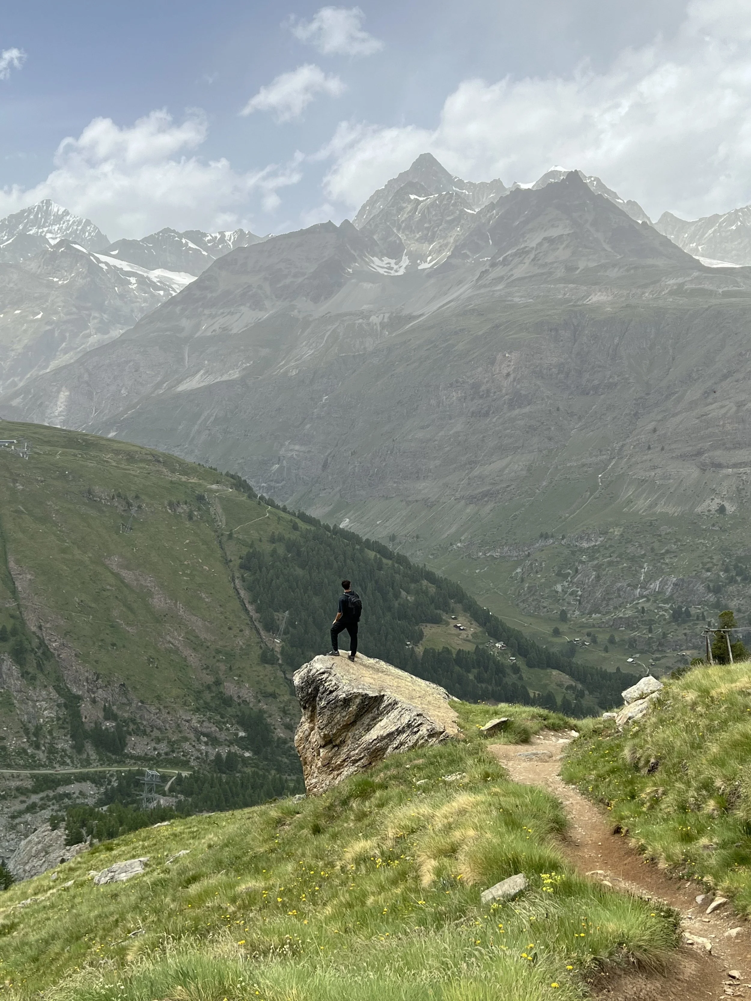 A person standing on a large rock ledge overlooking a mountain landscape with green hills, snow-capped peaks, and a partly cloudy sky.