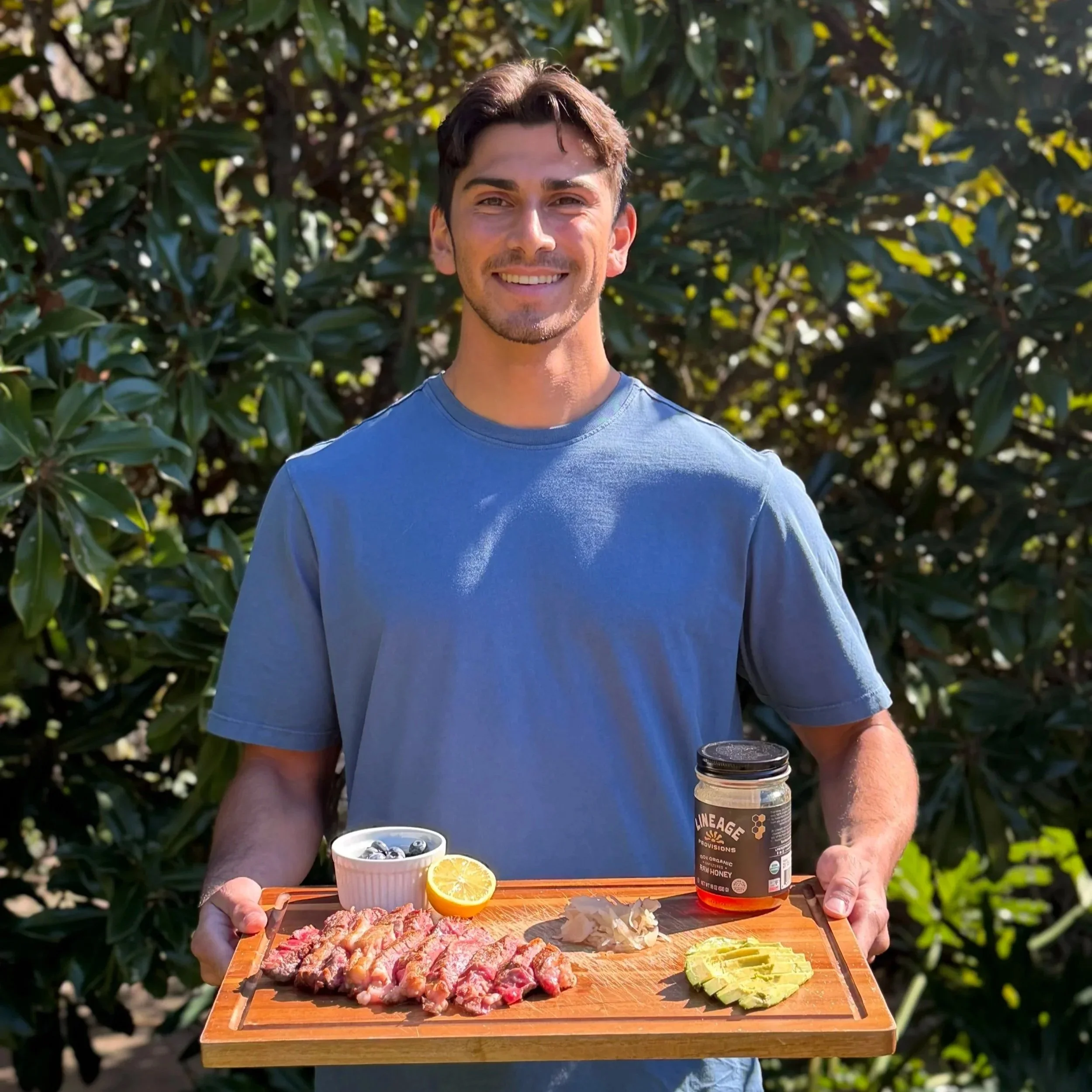A man holding a wooden tray with sliced steak, lemon, blueberries, honey, pickled ginger, and sliced avocado outdoors.