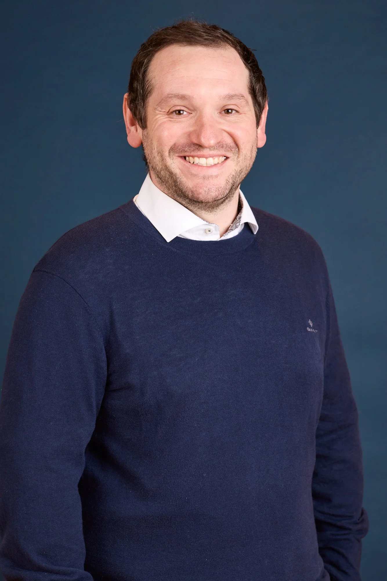 Ein lächelnder Mann mit kurzen braunen Haaren, im weißen Hemd und dunkelblauem Pullover vor einem blauen Hintergrund.