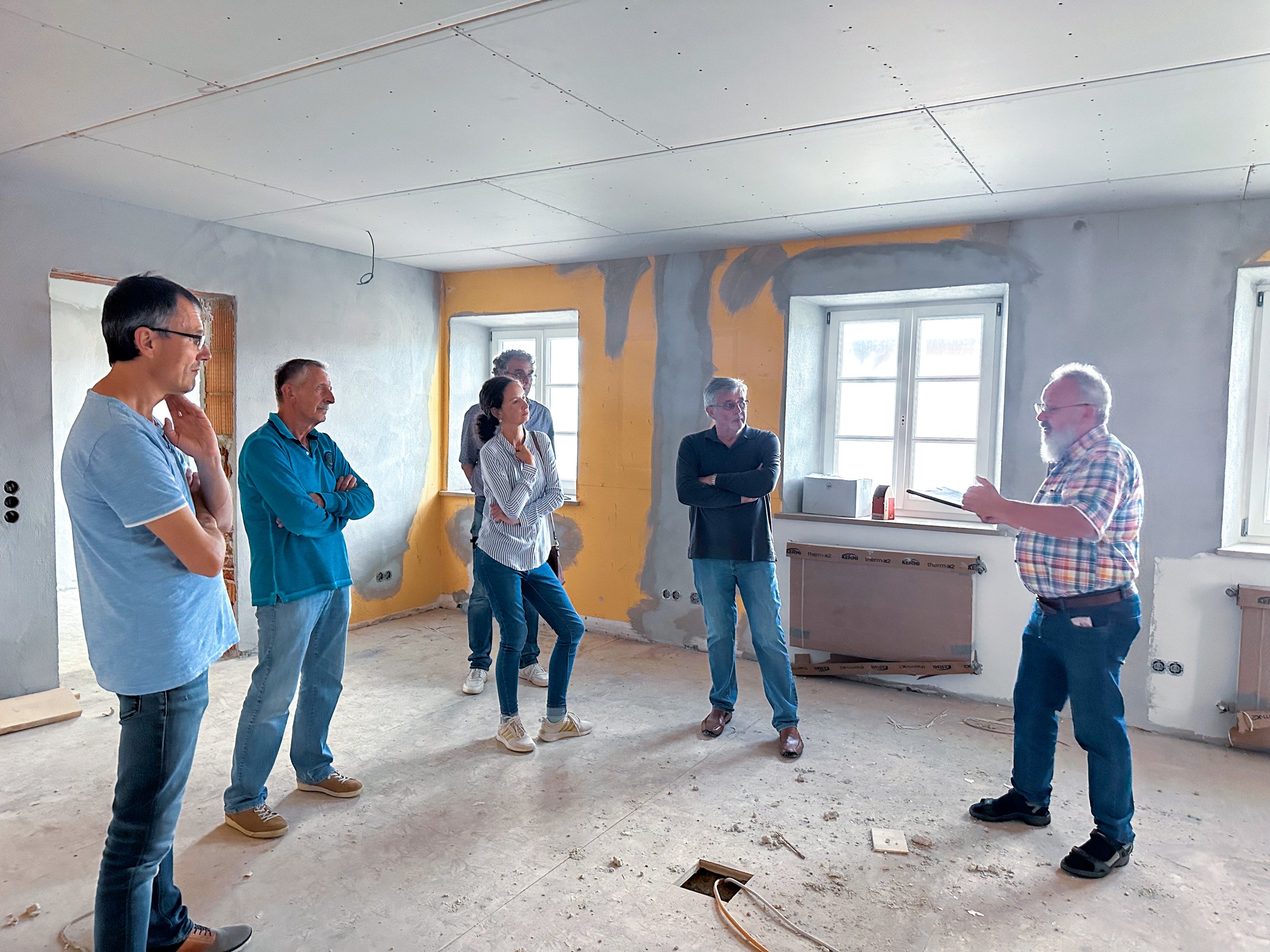 Eine Gruppe von sechs Menschen steht im Raum einer renovierten, leeren Wohnung, während ein Mann auf der rechten Seite mit einem Tablet spricht.