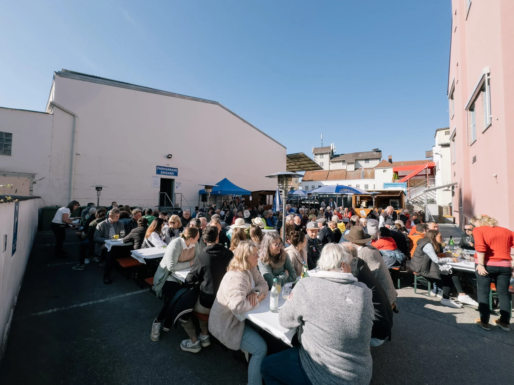 Verschiedene Menschen sitzen an Tischen im Freien vor einem Gebäude mit einer blauen Markise und einem Pavillon, bei schönem Wetter.