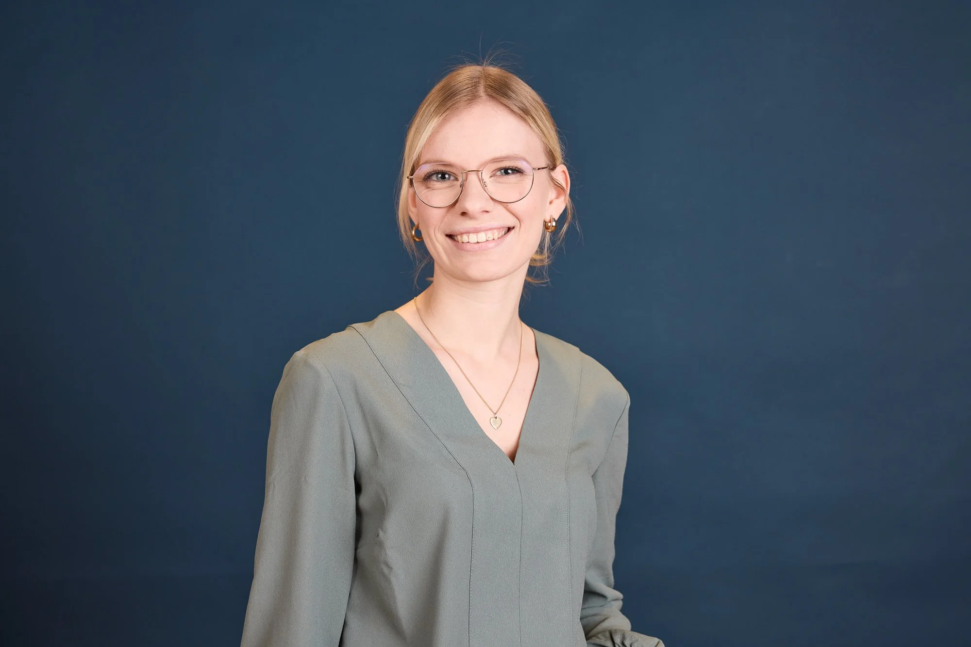 Junge Frau mit blonden Haaren, Brille, und Ohrringen vor dunklem blauen Hintergrund. Sie trägt eine graue Bluse und Halskette, lächelt und schaut zur Seite.