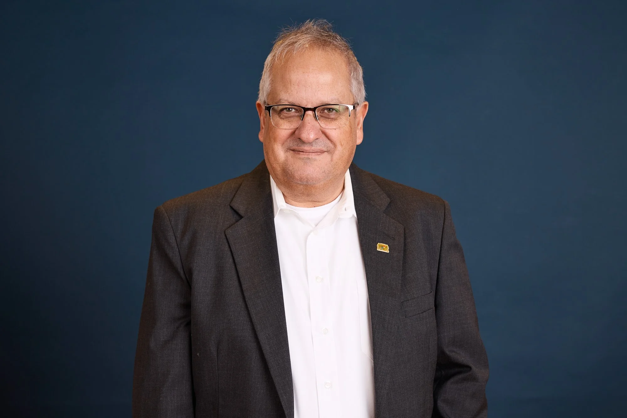 Ein Mann trägt eine dunkle Anzugjacke und eine weiße Hemd, steht vor einem blauen Hintergrund, trägt eine Brille, hat kurzes graues Haar und lächelt leicht.