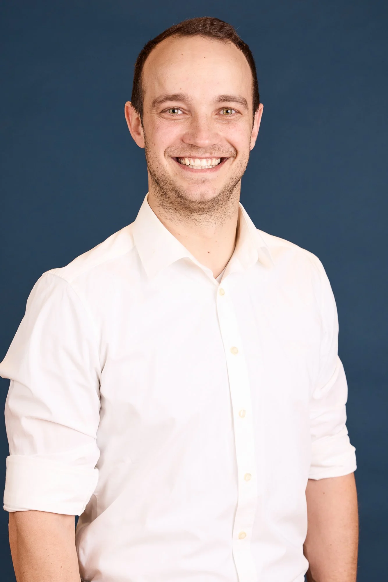 Ein lächelnder junger Mann trägt ein weißes Hemd gegen einen blauen Hintergrund.