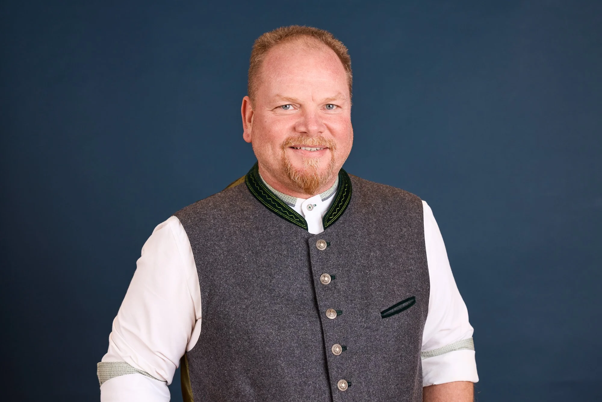 Mann in traditioneller deutscher Kleidung, lächelnd, vor dunklem Hintergrund.