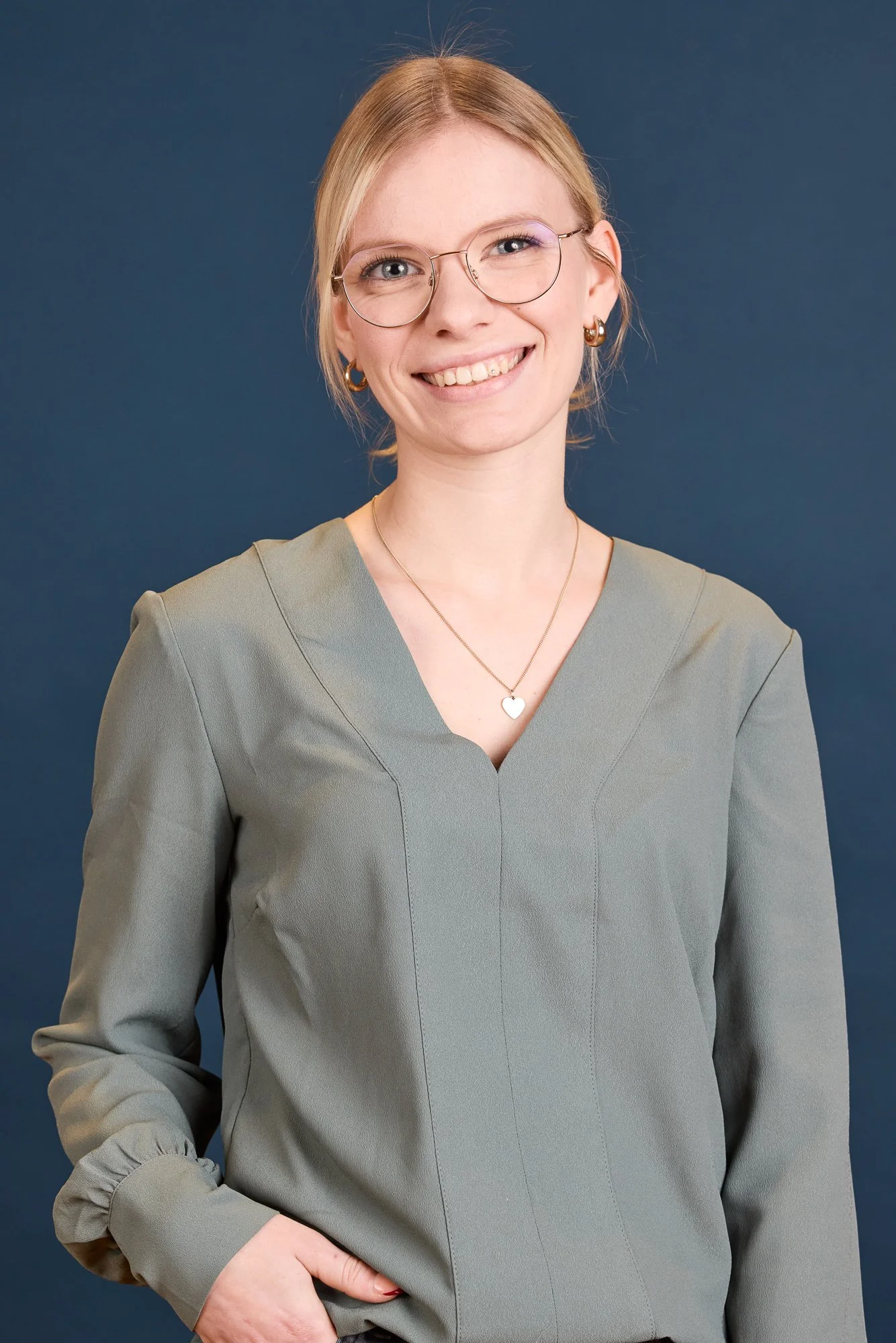 Porträt einer lachenden jungen Frau mit Brille, kurzen blonden Haaren und Ohrringen vor einem dunklen Hintergrund.