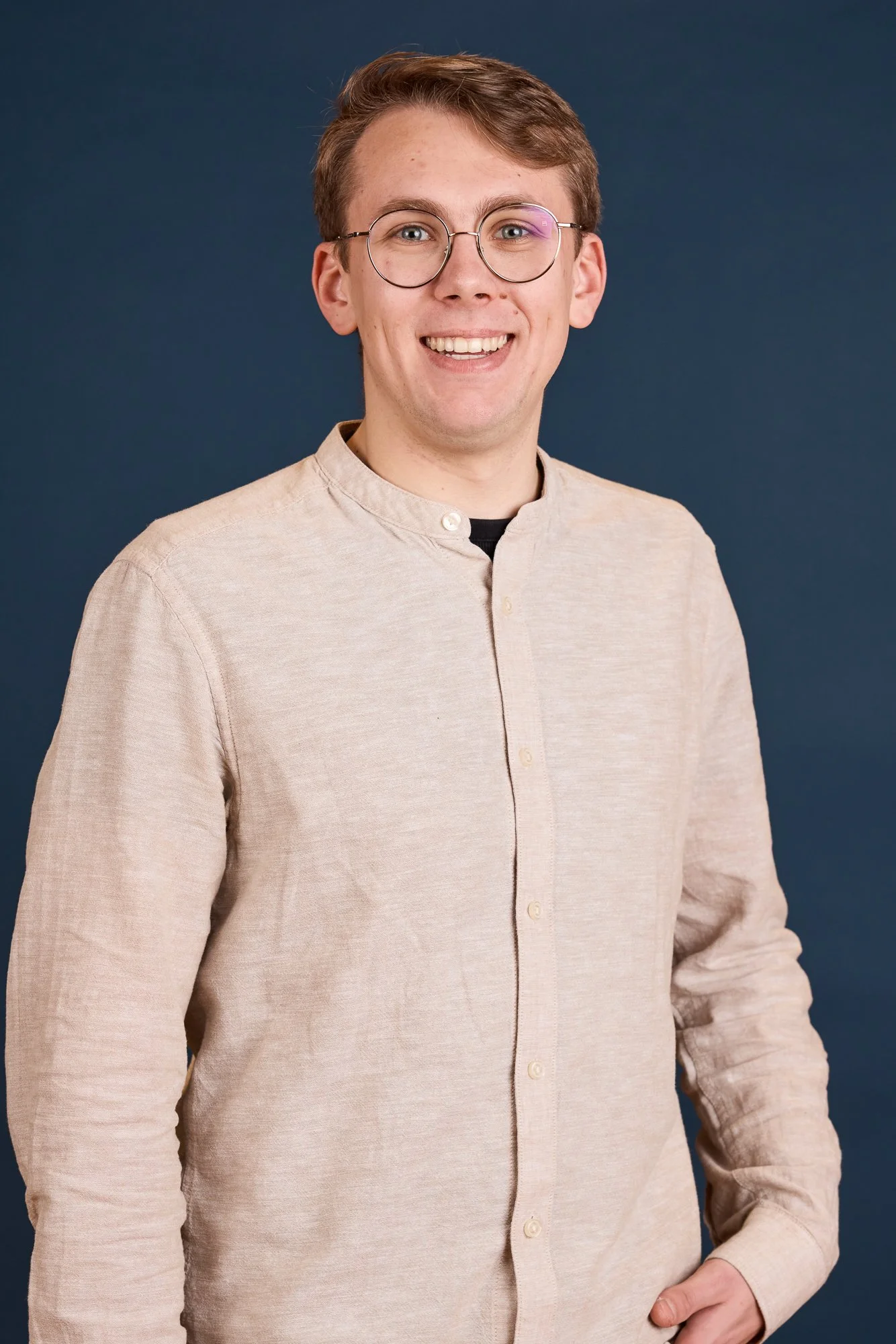 Ein junger Mann mit Brille, hellbeigem Hemd und braunen Haaren, lächelnd vor einem dunklen Hintergrund.