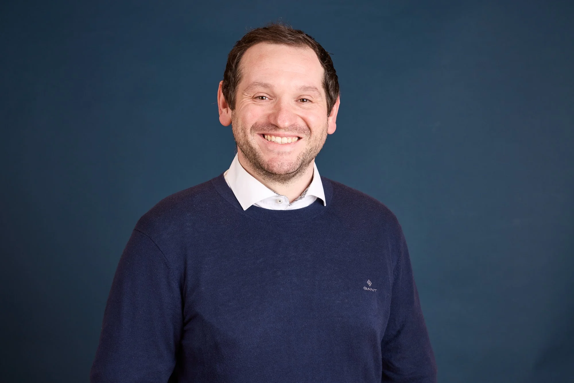 Ein Mann mit braunen Haaren, Bart und einem Lächeln trägt einen dunkelblauen Pullover und ein weißes Hemd vor einem dunkelblauen Hintergrund.