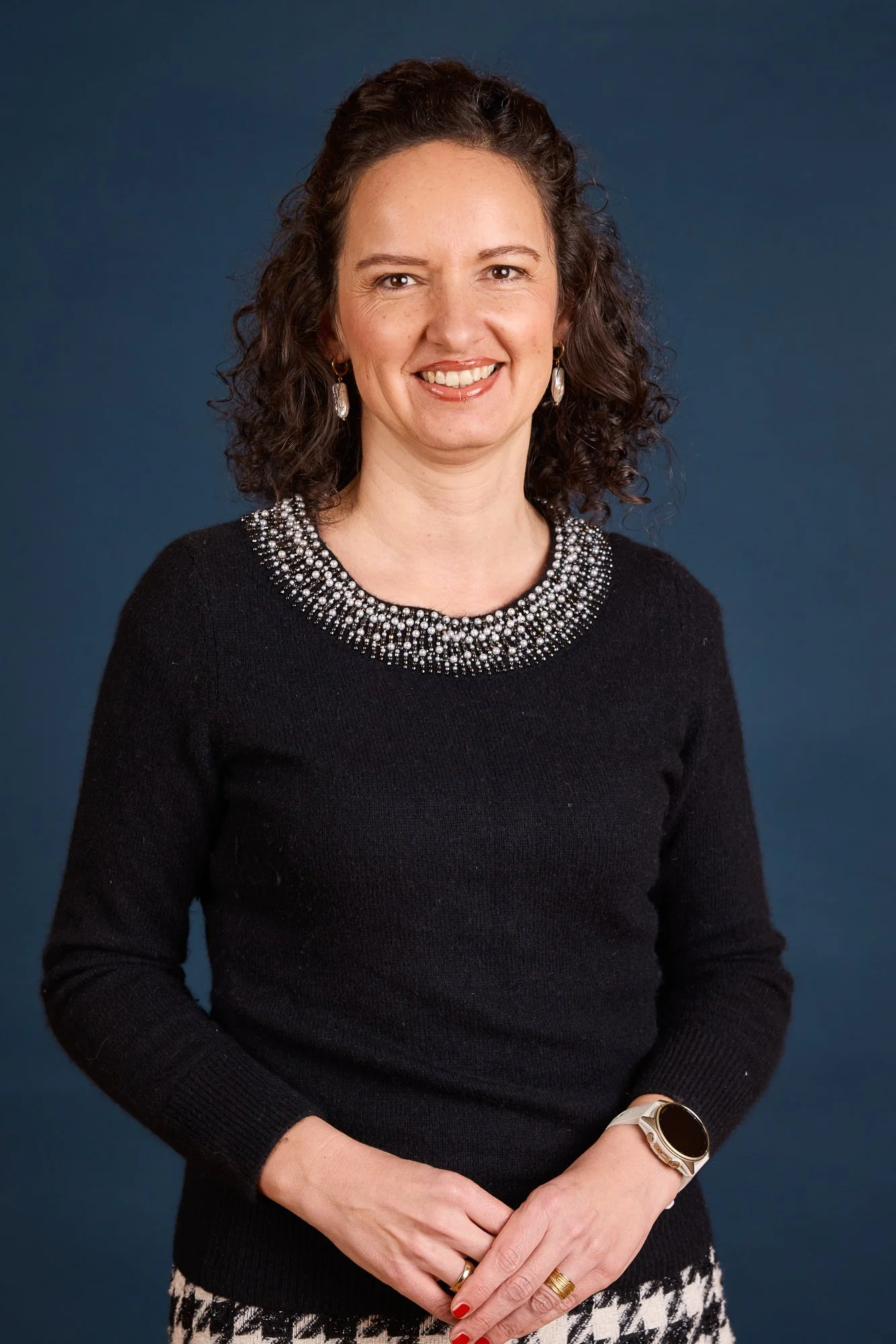Eine lachende Frau mit dunklen, lockigen Haaren, trägt eine schwarze Strickjacke mit Perlenverzierung am Ausschnitt, eine Armbanduhr, Ringe und Ohrringe, steht vor einem blauen Hintergrund.