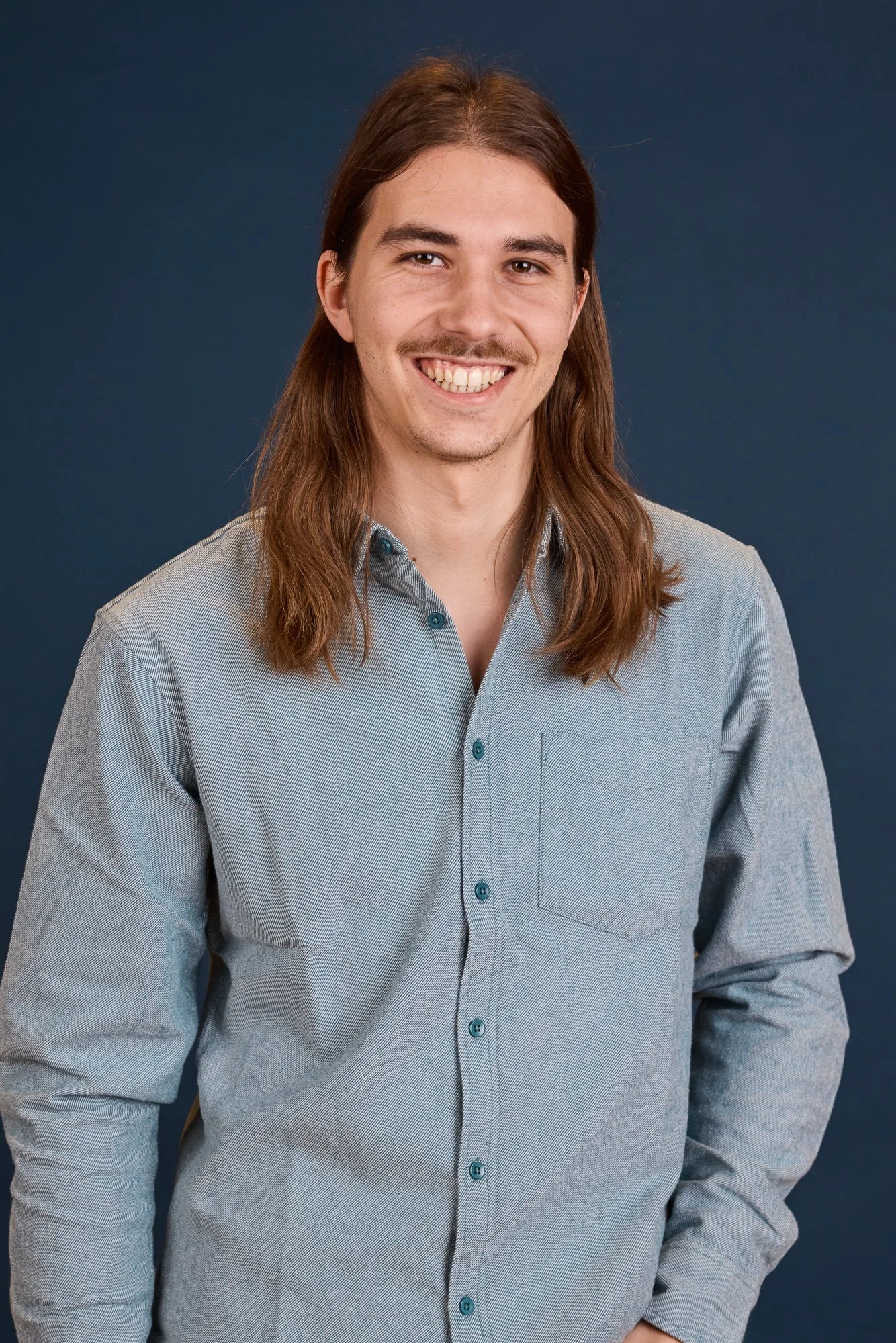 Mann mit langen braunen Haaren, Bart, grauem Hemd vor dunkelblauem Hintergrund.