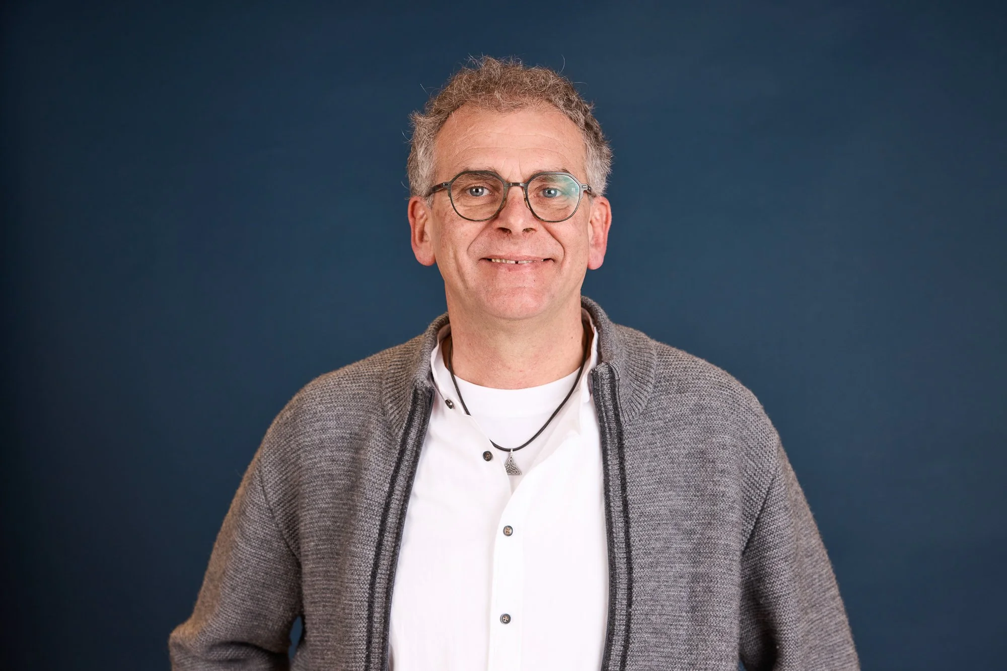 Mann mit Brille, grauem Handwerkerpullover und weißem Hemd, vor dunkelblauem Hintergrund.