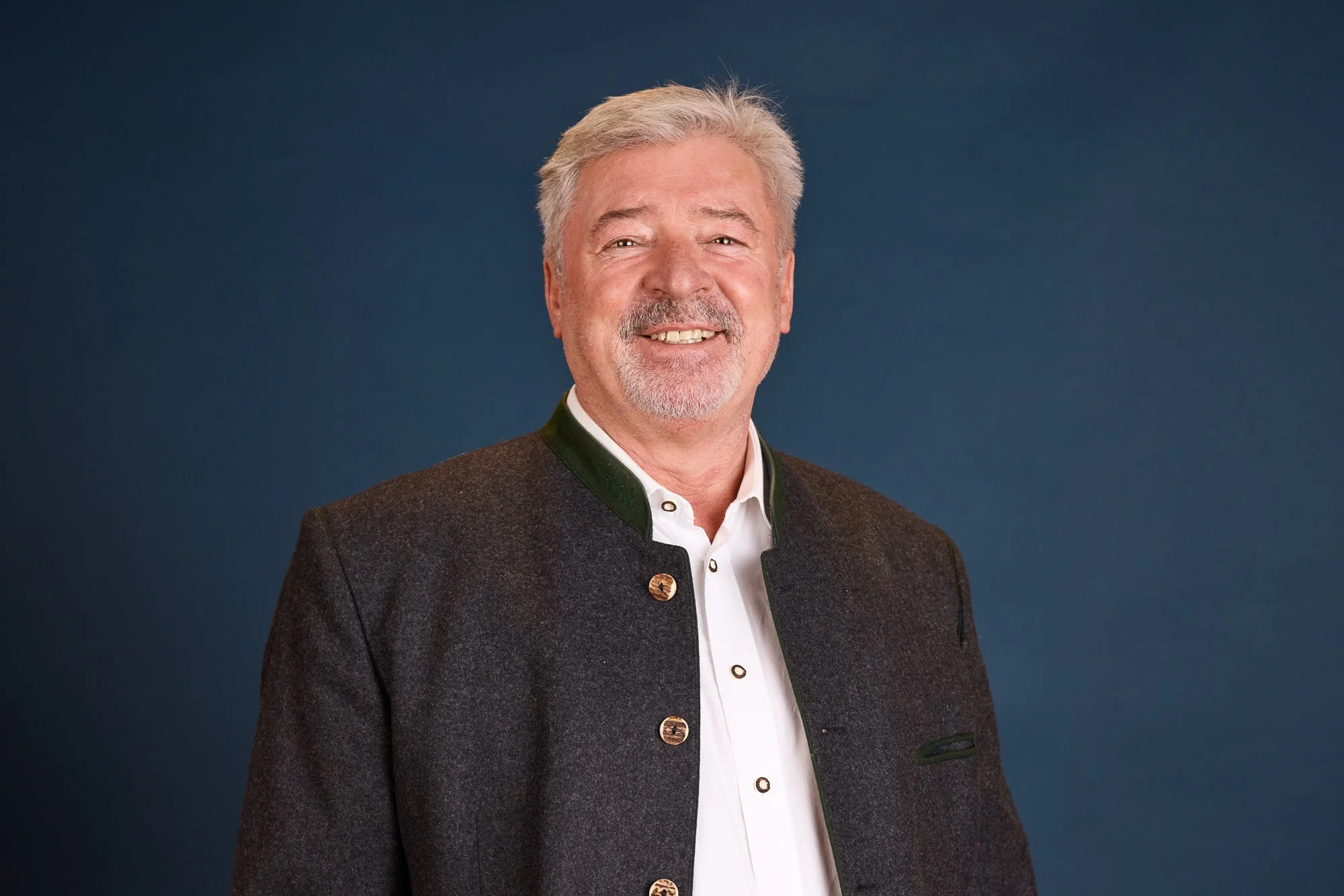 Ein älterer Mann mit grauem Haar und Bart, trägt eine traditionelle dunkle Trachtenjacke mit grünem Kragen, vor einem dunklen Hintergrund.