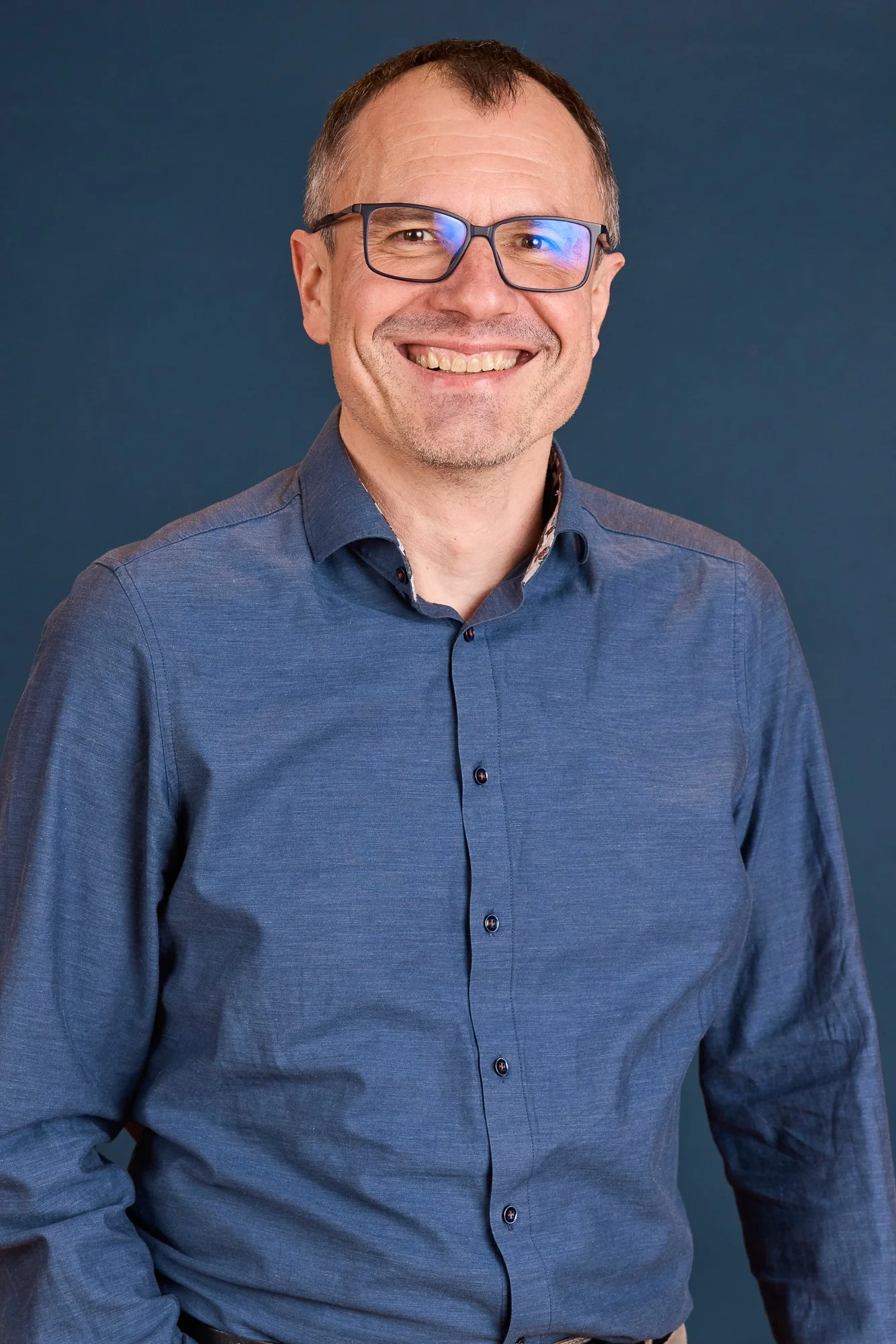 Ein Mann trägt eine blaue Hemd und trägt eine Brille, lächelt freundlich, vor einem dunklen Hintergrund.