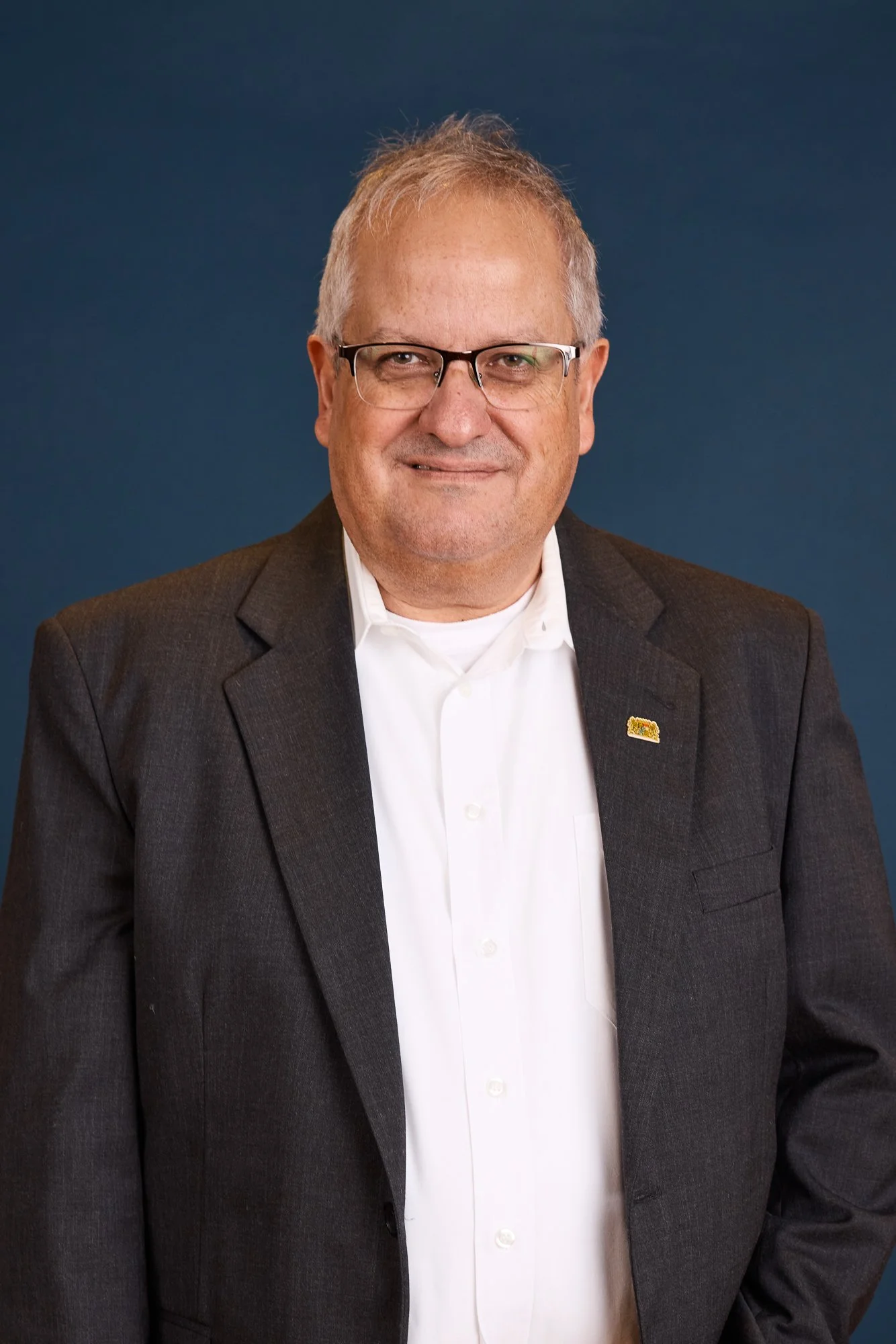 Ein älterer Mann mit Brille, grauem Haar und einem schwarzen Anzug steht vor einem blauen Hintergrund.