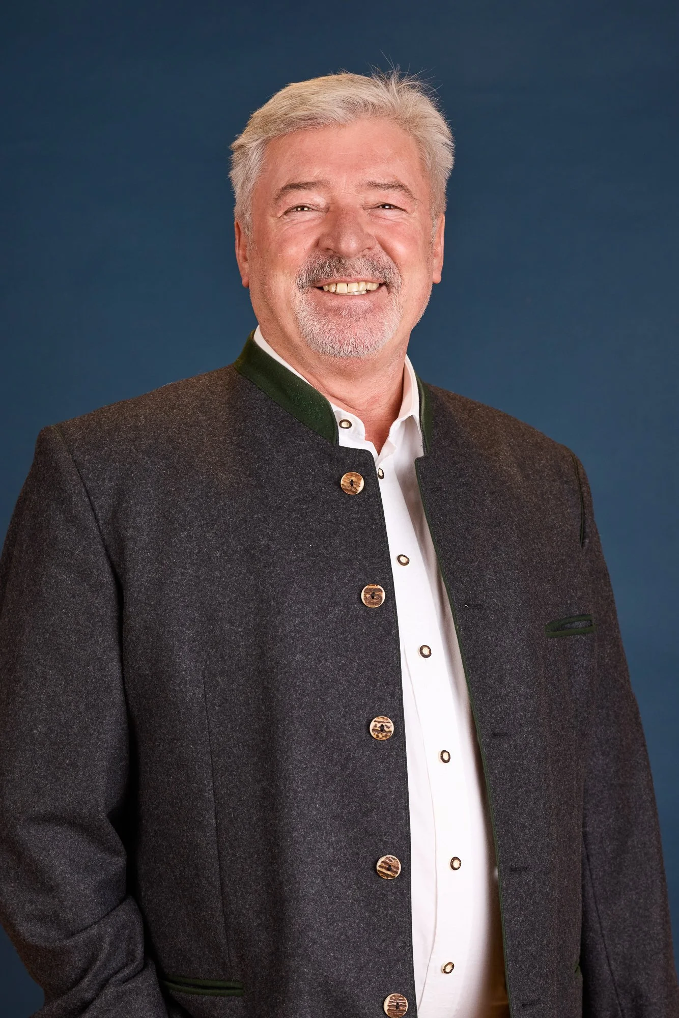 Ein älterer Mann mit grauem Haar und Bart lächelt. Er trägt eine traditionelle dunkle Jacke mit goldenen Knöpfen und ein weißes Hemd. Der Hintergrund ist blau.