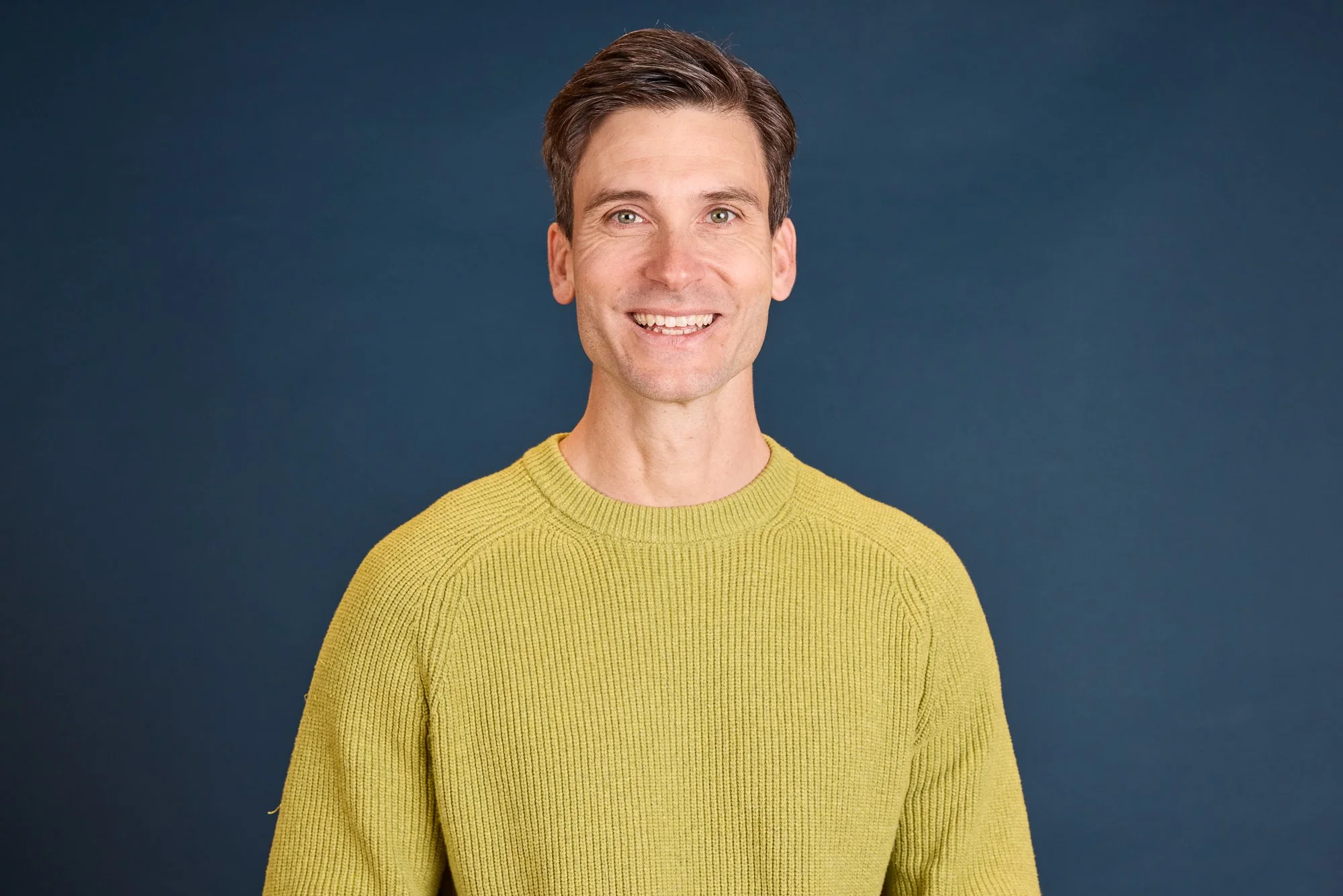 Ein Mann mit braunen Haaren trägt einen gelben Pullover vor einem blauen Hintergrund und lächelt in die Kamera.