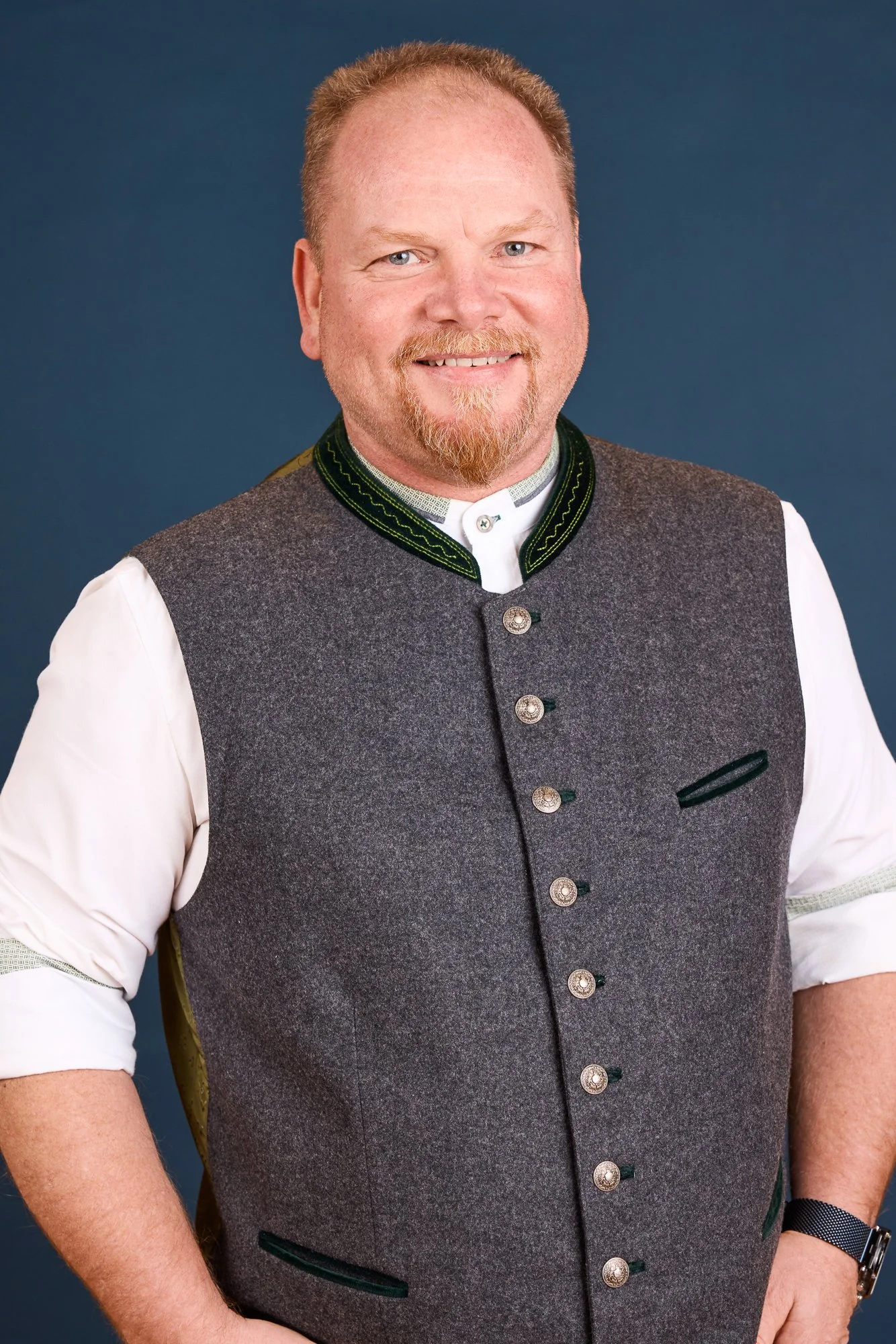 Mann trägt traditionelle bayerische Tracht, bestehend aus Weste und Hemd, vor einem blauen Hintergrund.