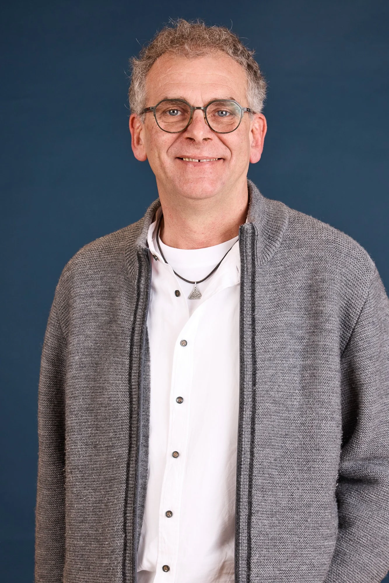 Ein älterer Mann mit Brille, grauem Shorts, weißem Hemd und grauem Pullover vor dunklem Hintergrund.