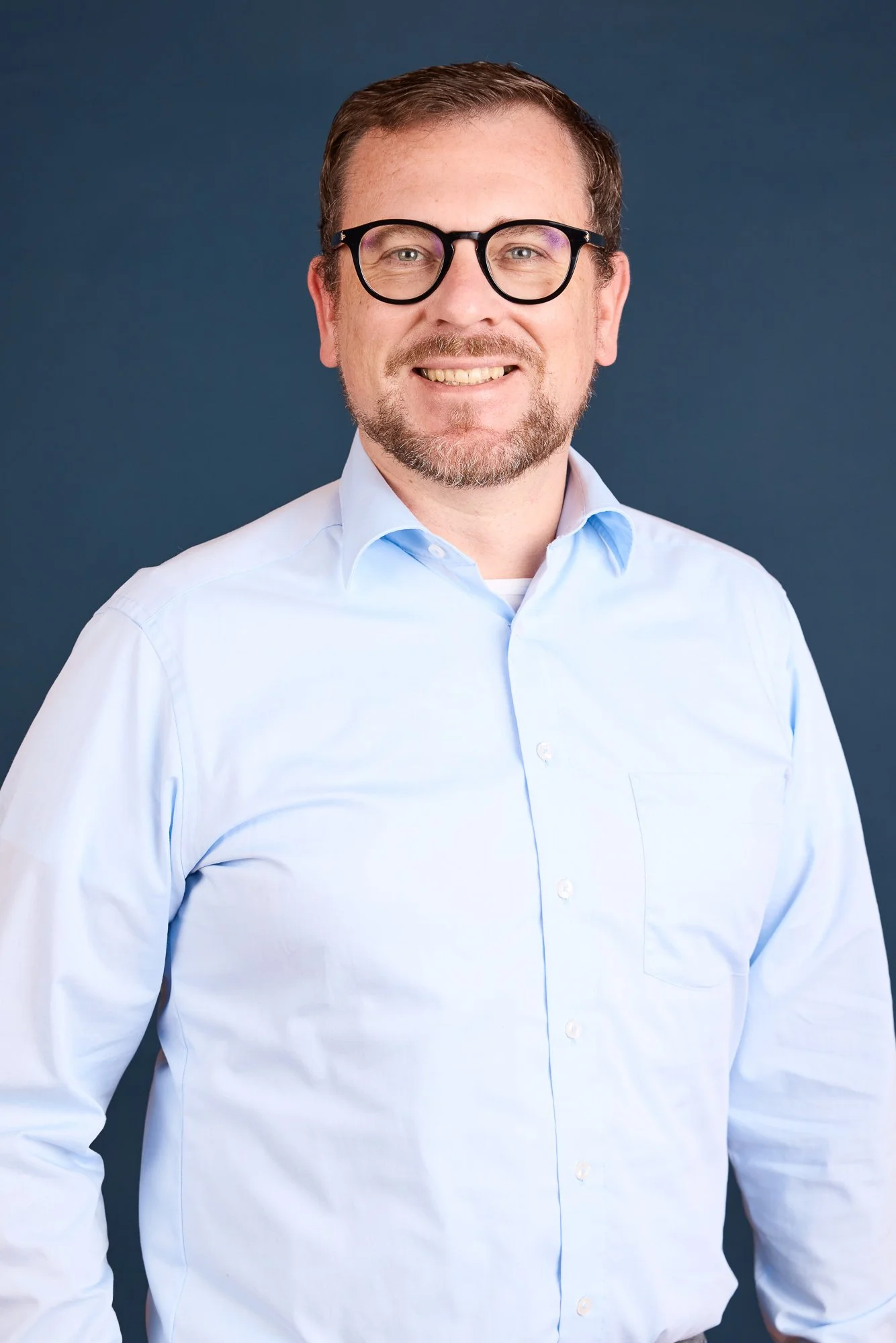Porträt eines Mannes mit Brille in einem weißen Hemd vor blauem Hintergrund.