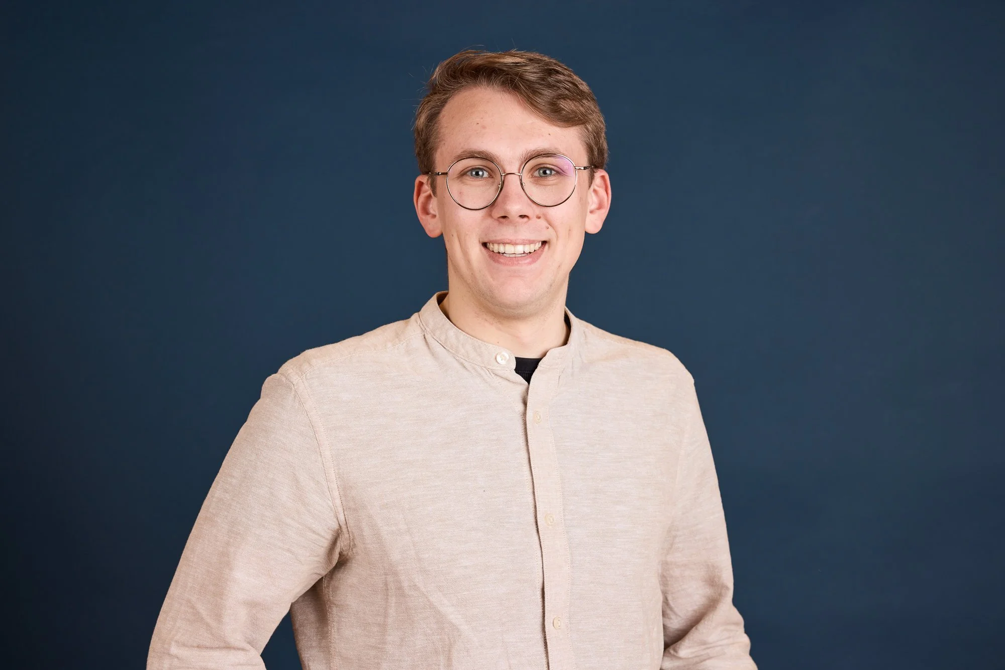 Junger Mann mit Brille vor dunkelblauem Hintergrund, lächelnd und casual gekleidet.