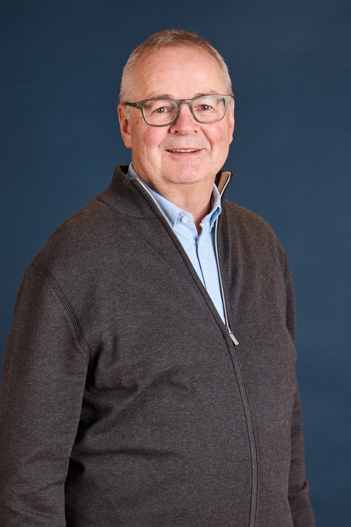Ein älterer Mann mit Brille, dunkelbraunem Pullover und blauer Hemd, lächelt vor einem dunklen blauen Hintergrund.