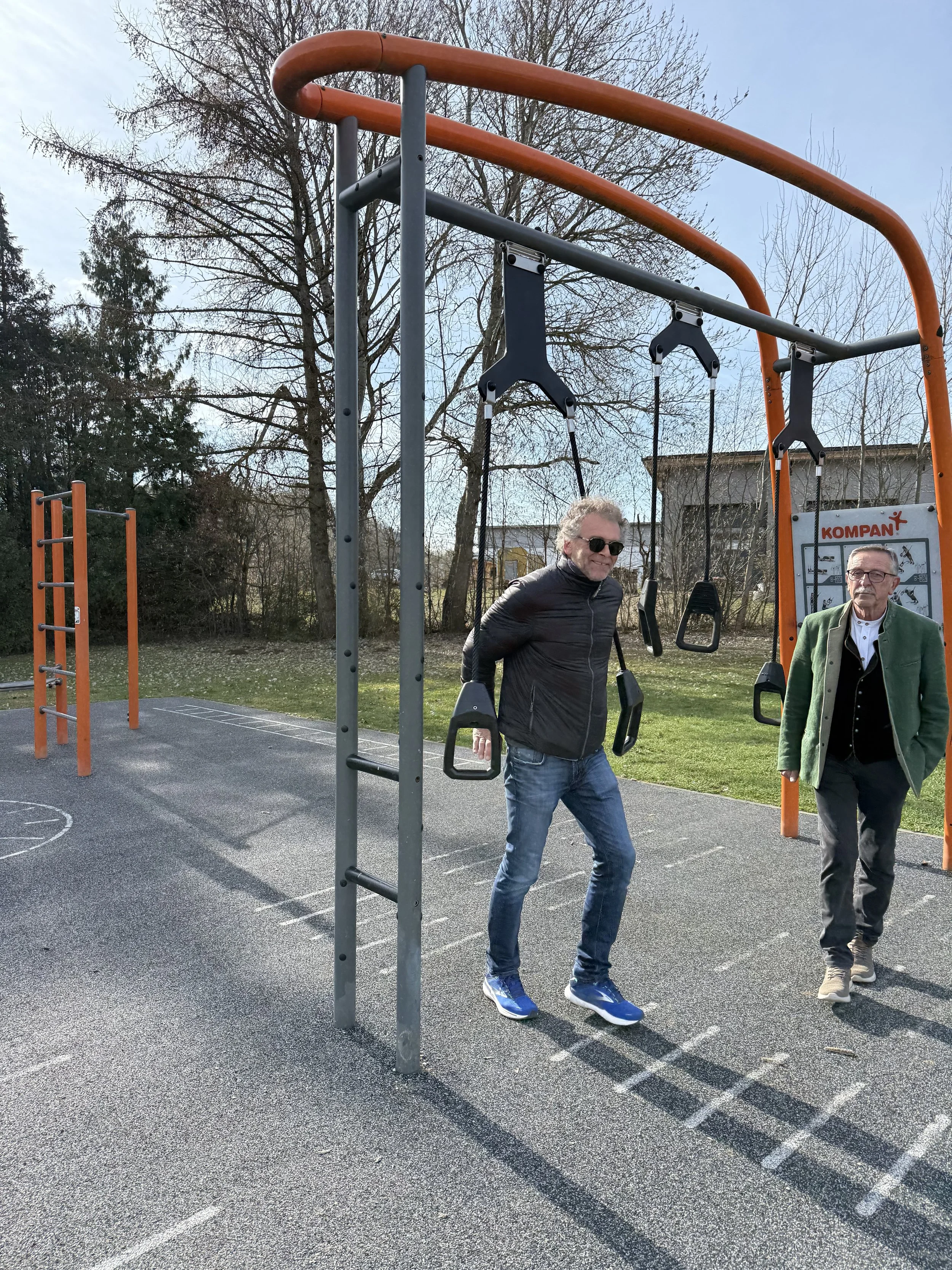 Zwei Männer stehen unter einer Schaukel im Freien auf einem Spielplatz. Es ist Tag und die Sonne scheint, im Hintergrund sind Bäume und ein Gebäude sichtbar.