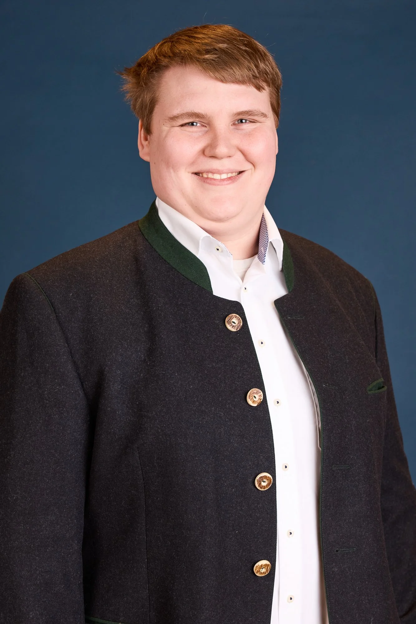 Junge Mann in traditionellen schwarzen Trachtenjacke mit weißen Hemd vor blauem Hintergrund.