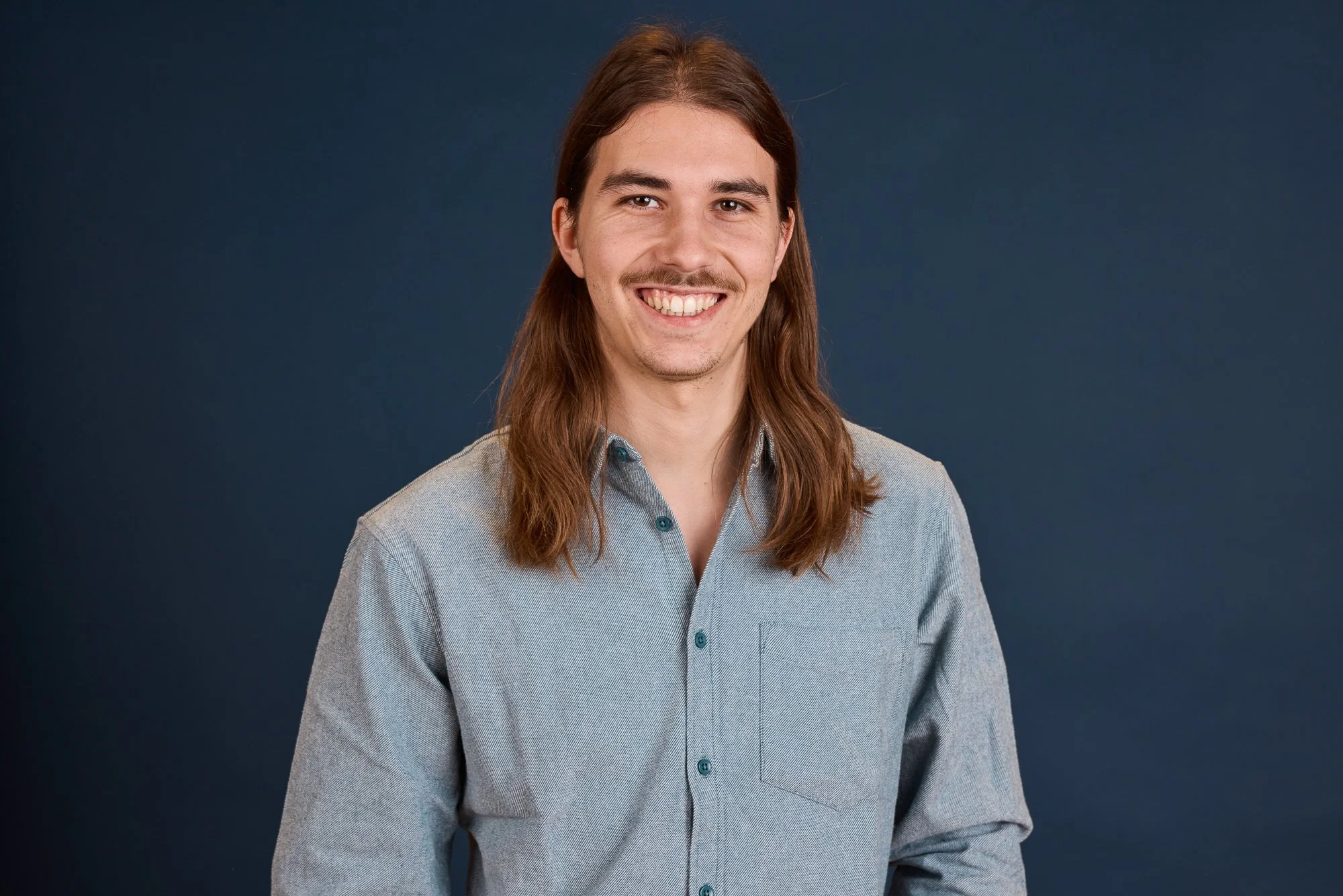 Ein Lächelnder Mann mit langen braunen Haaren und einem hellgrauen Hemd vor einem dunkelblauen Hintergrund.