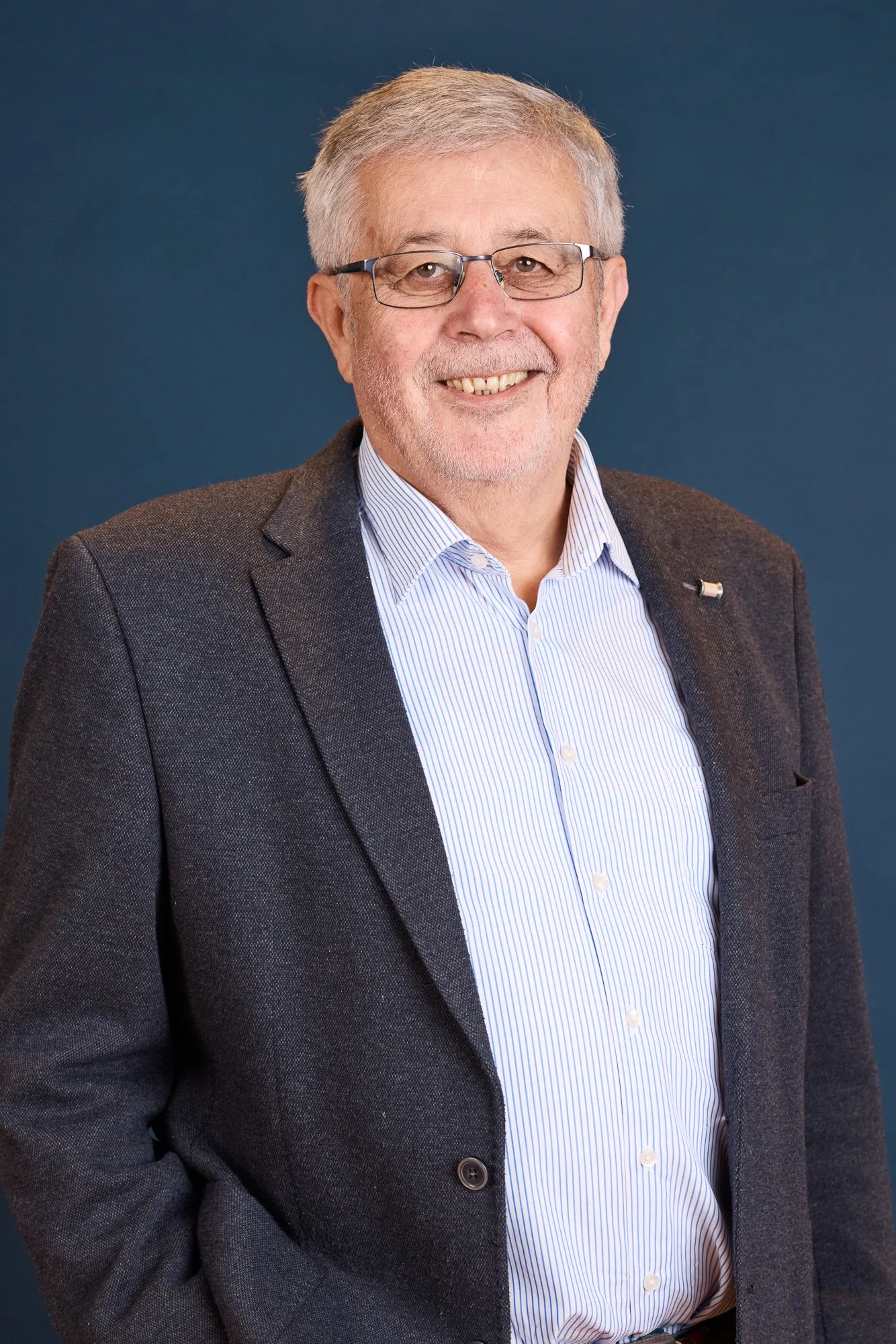 Ein älterer Mann trägt eine Brille, ein dunkelgraues Jackett und ein weiß-blau gestreiftes Hemd, und lächelt vor einem dunklen blauen Hintergrund.