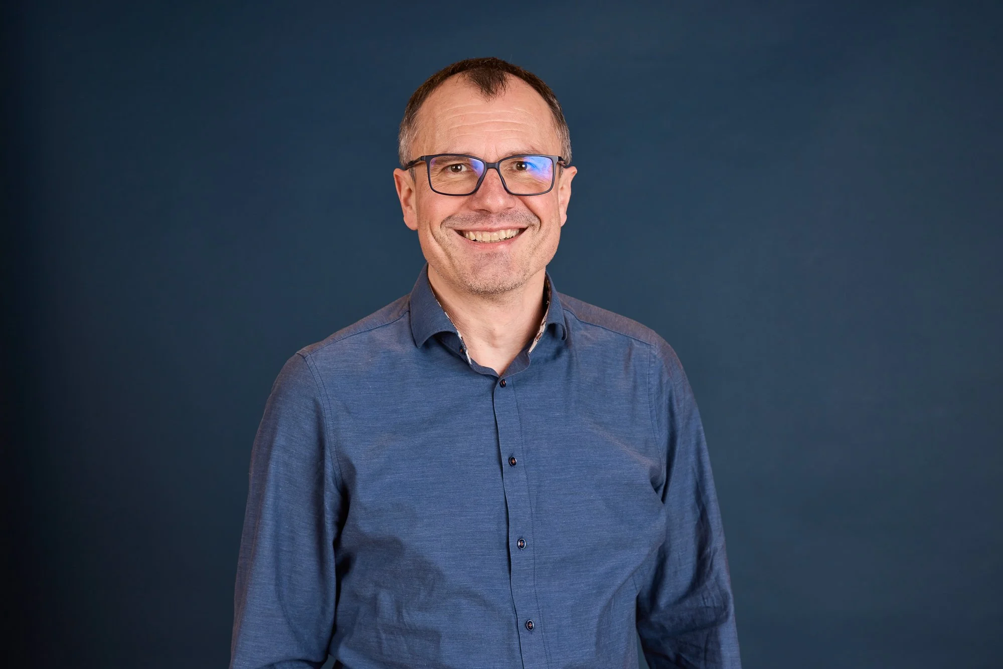 Ein lachender Mann trägt eine Brille und ein dunkelblaues Hemd vor einem dunklen Hintergrund.