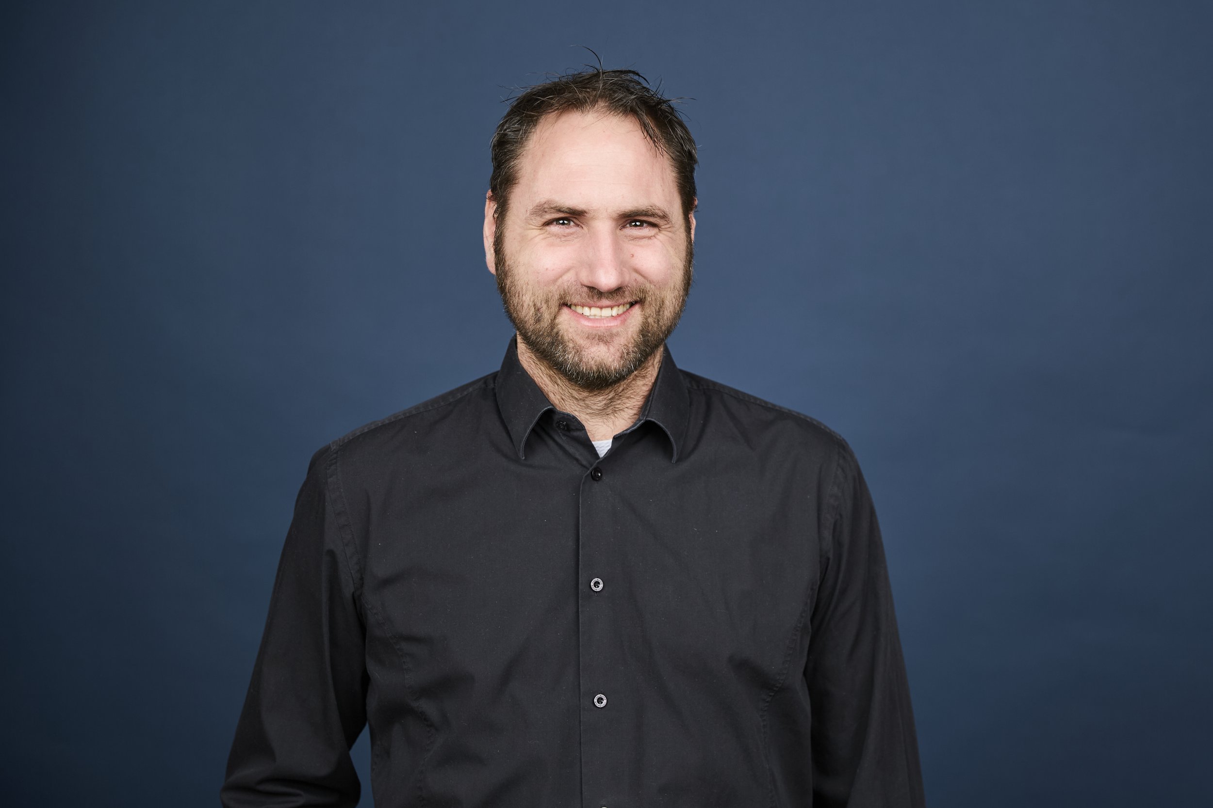 Ein Mann mit braunem Haar, Bart und einem schwarzen Hemd, lächelnd vor einem dunklen blauen Hintergrund.