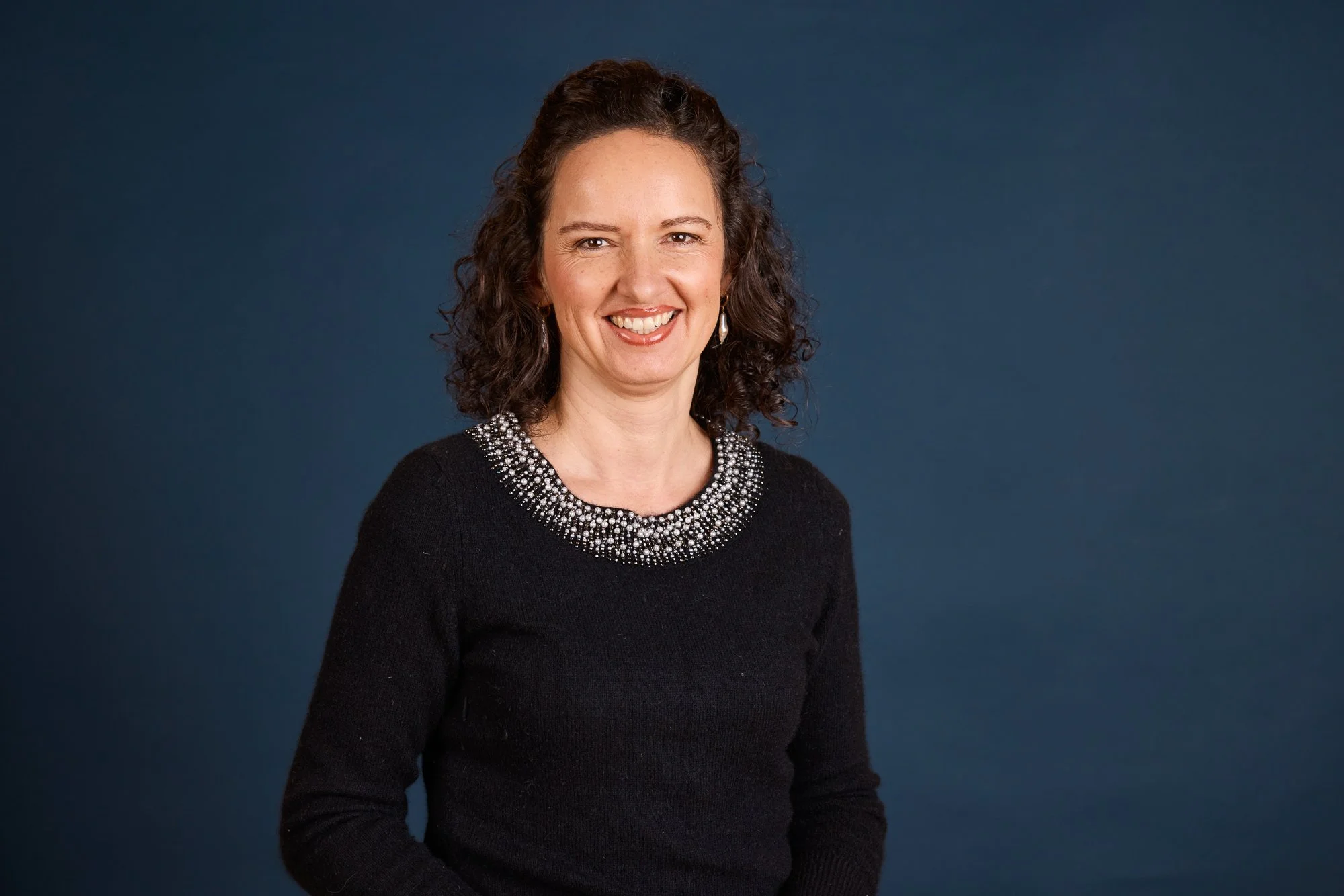 Eine lächelnde Frau mit braunen, lockigen Haaren, die ein schwarzes Oberteil mit einem verzierten Halsausschnitt trägt, vor einem dunklen blauen Hintergrund.