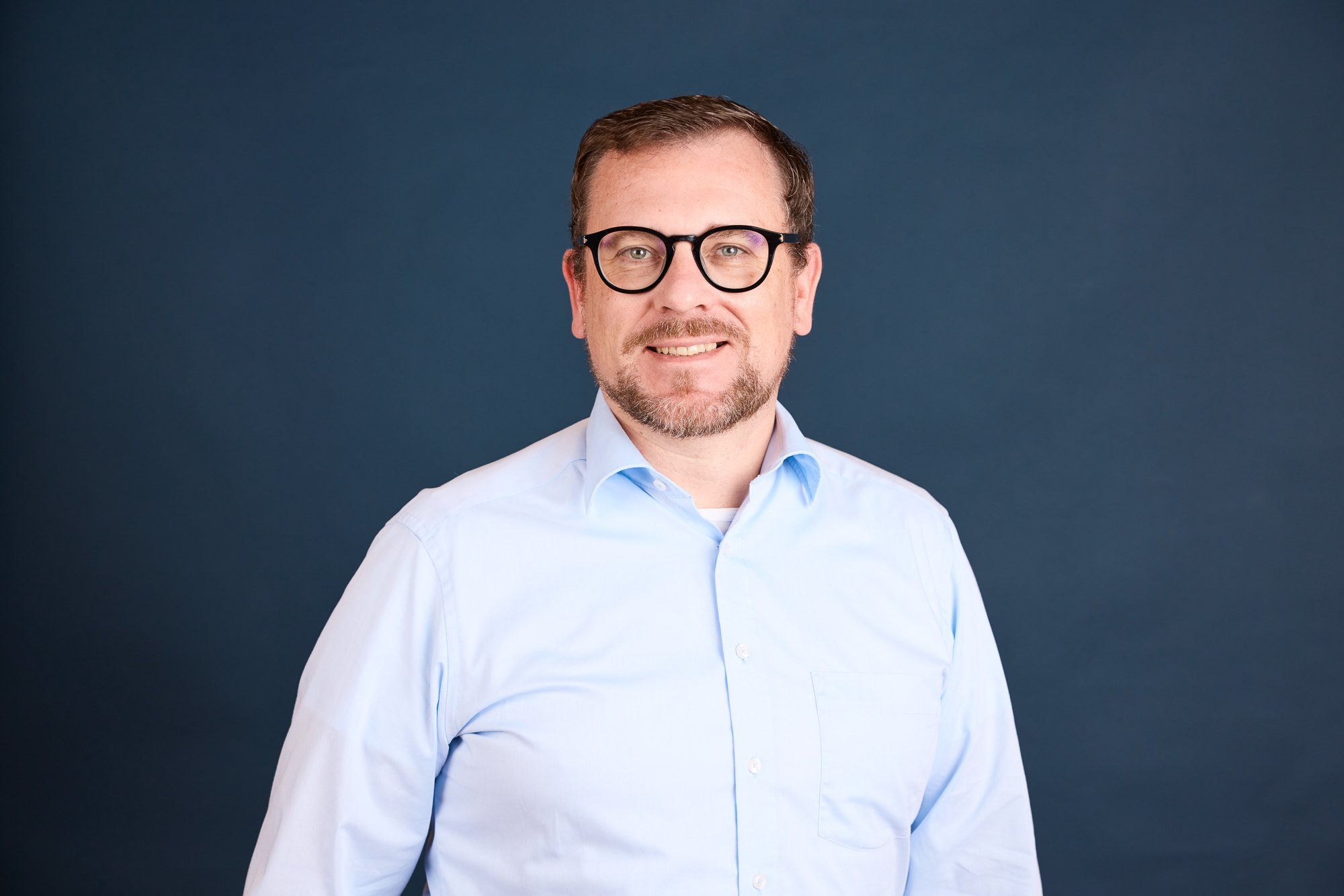Porträt eines Mannes mit Brille, Bart und Kurzhaar, trägt ein hellblaues Hemd, vor dunklem blauen Hintergrund.