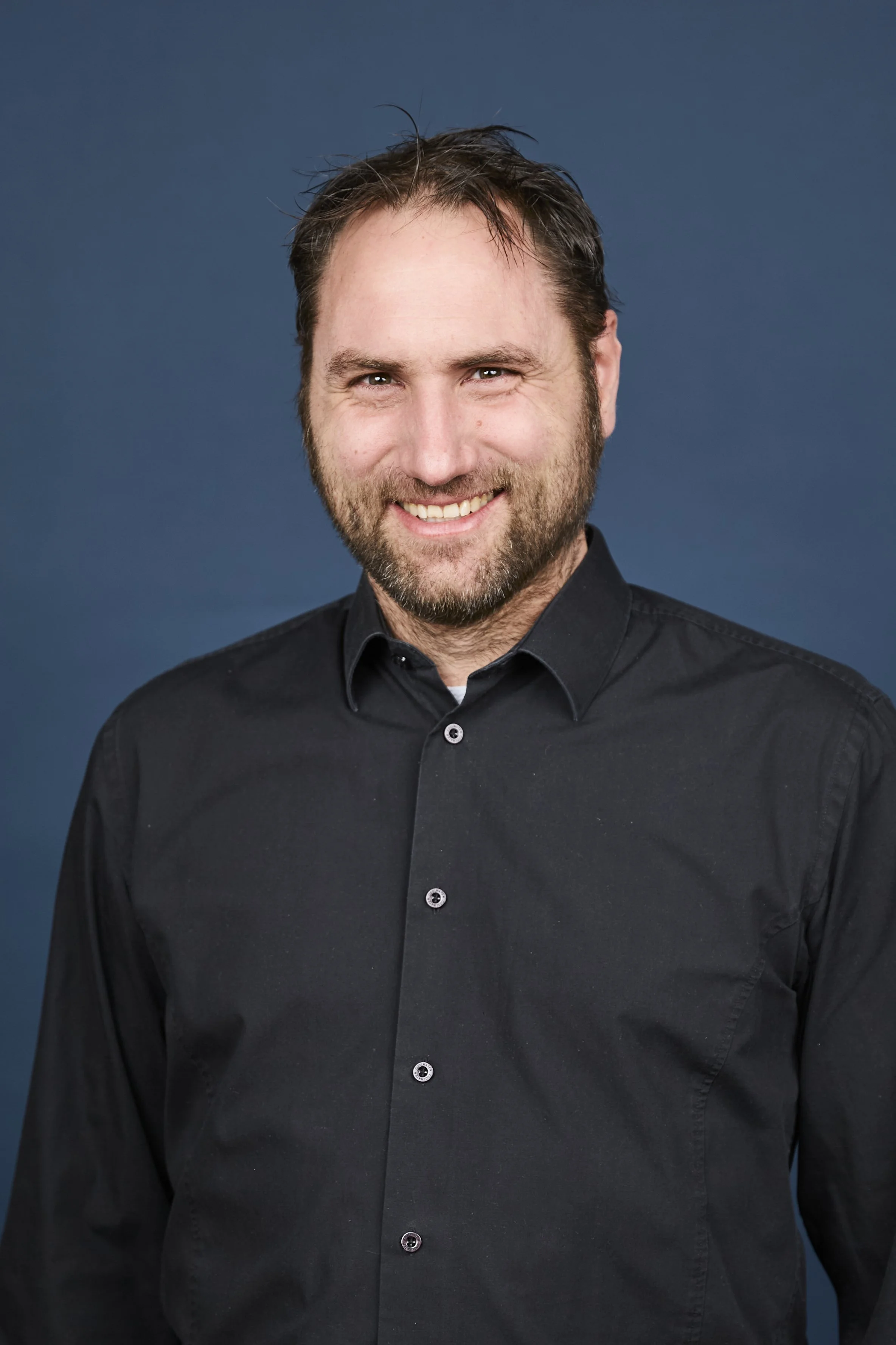 Ein Mann mit braunem Haar, Bart und einem schwarzen Hemd vor dunkelblauem Hintergrund, lächelnd in die Kamera schauend.