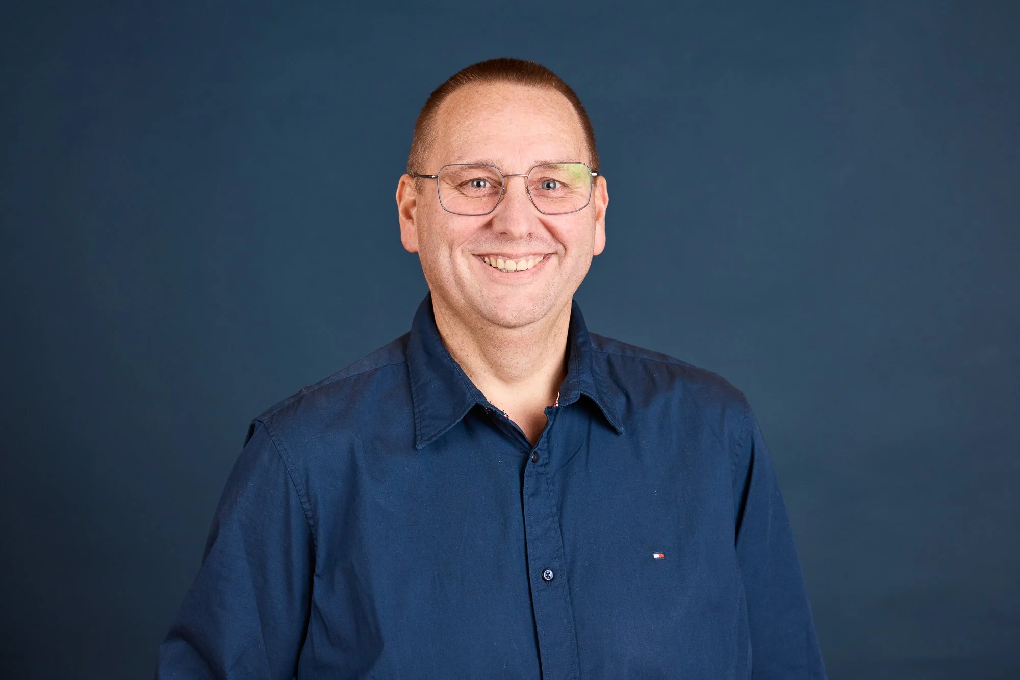 Ein lächelnder Mann mit Brille, kurzem Haar und einem dunkelblauen Hemd vor einem dunklen Hintergrund.