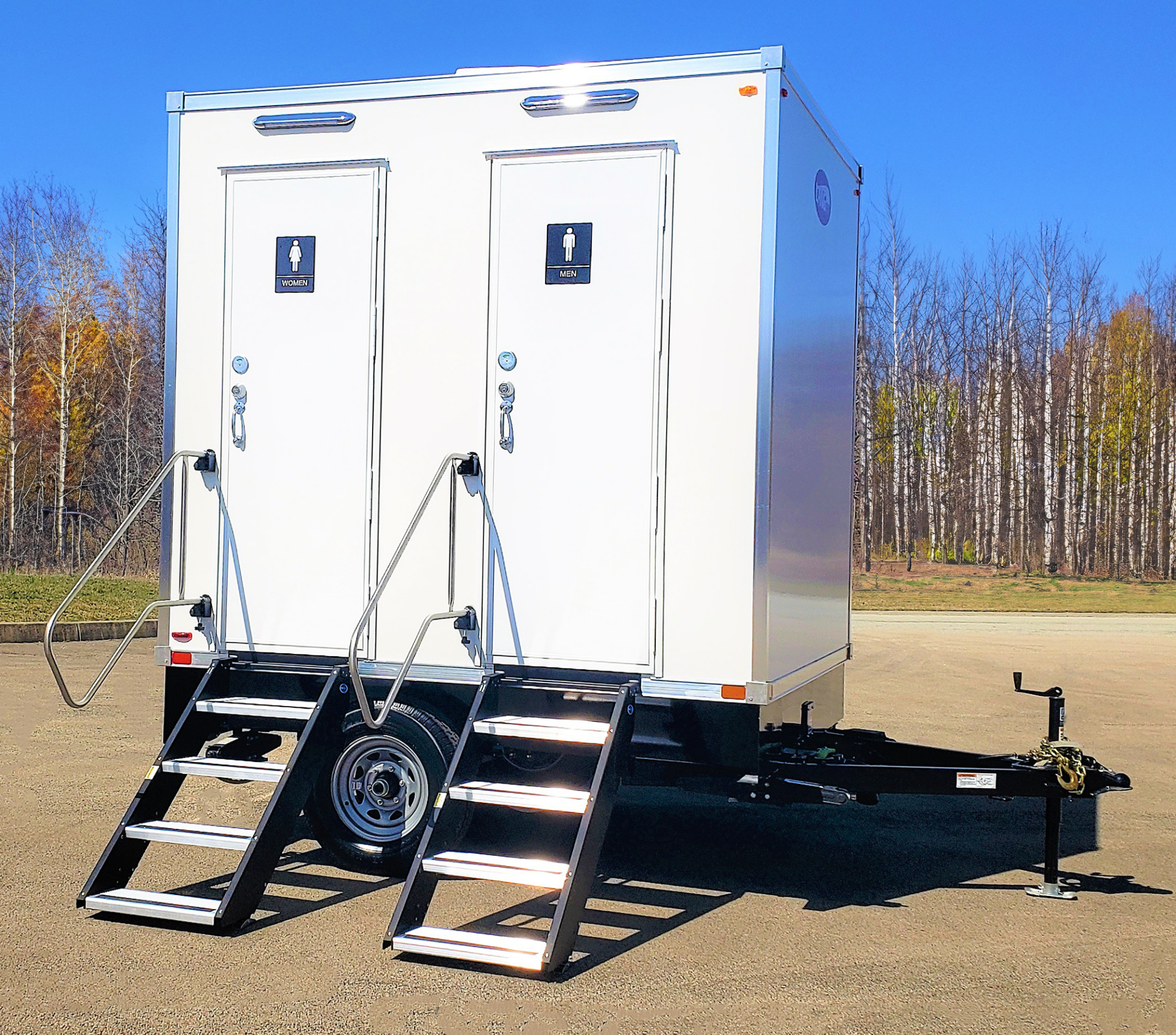 White mobile restroom trailer with separate facilities for women and men, two external steps, metal handrails, and a tongue for hitching to a vehicle, set on a flat area with trees in the background.