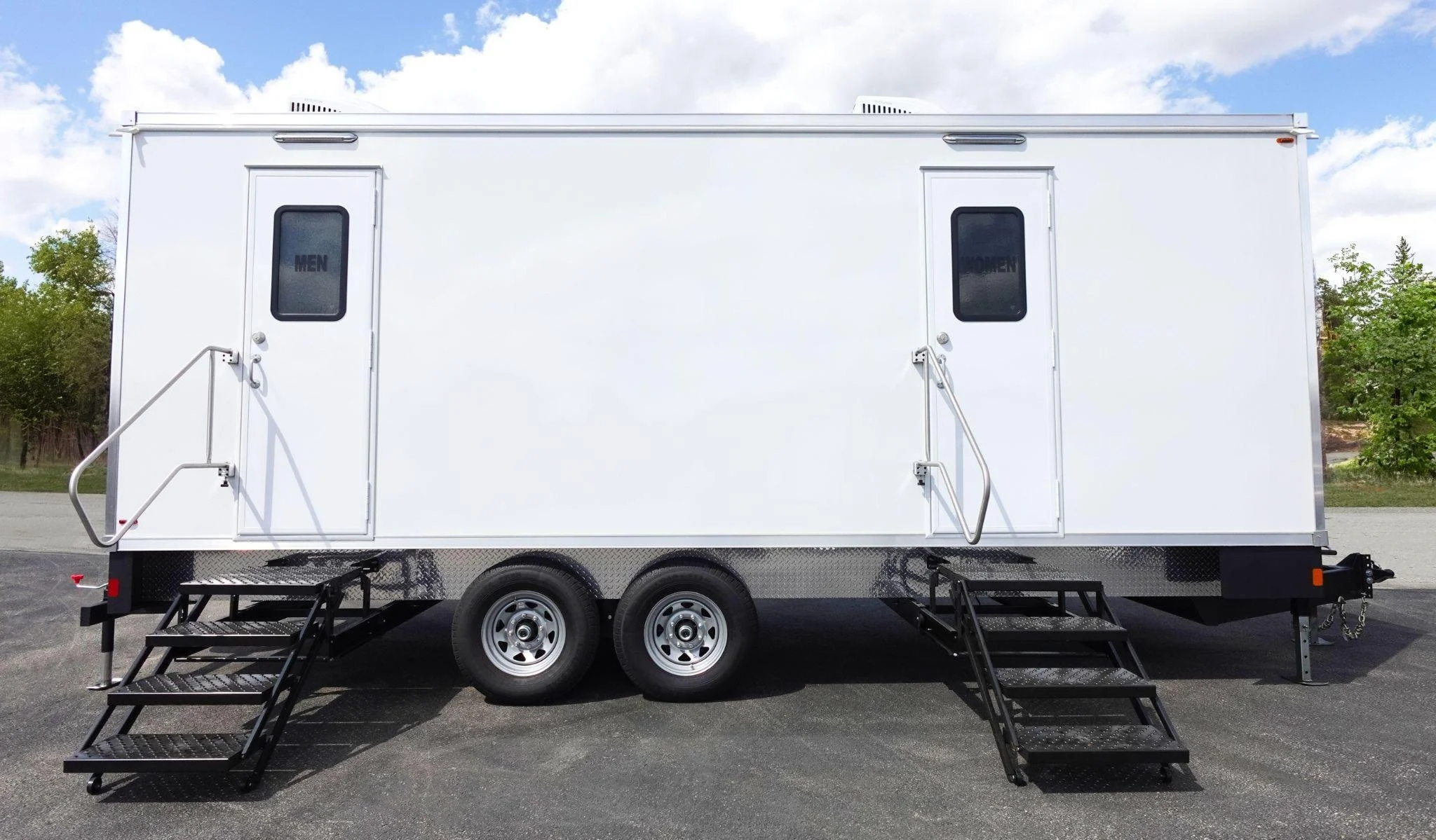 A white mobile restroom trailer with two doors labeled 'Men' and 'Women', black metal stairs, and black handrails, parked on pavement with trees and blue sky in the background.