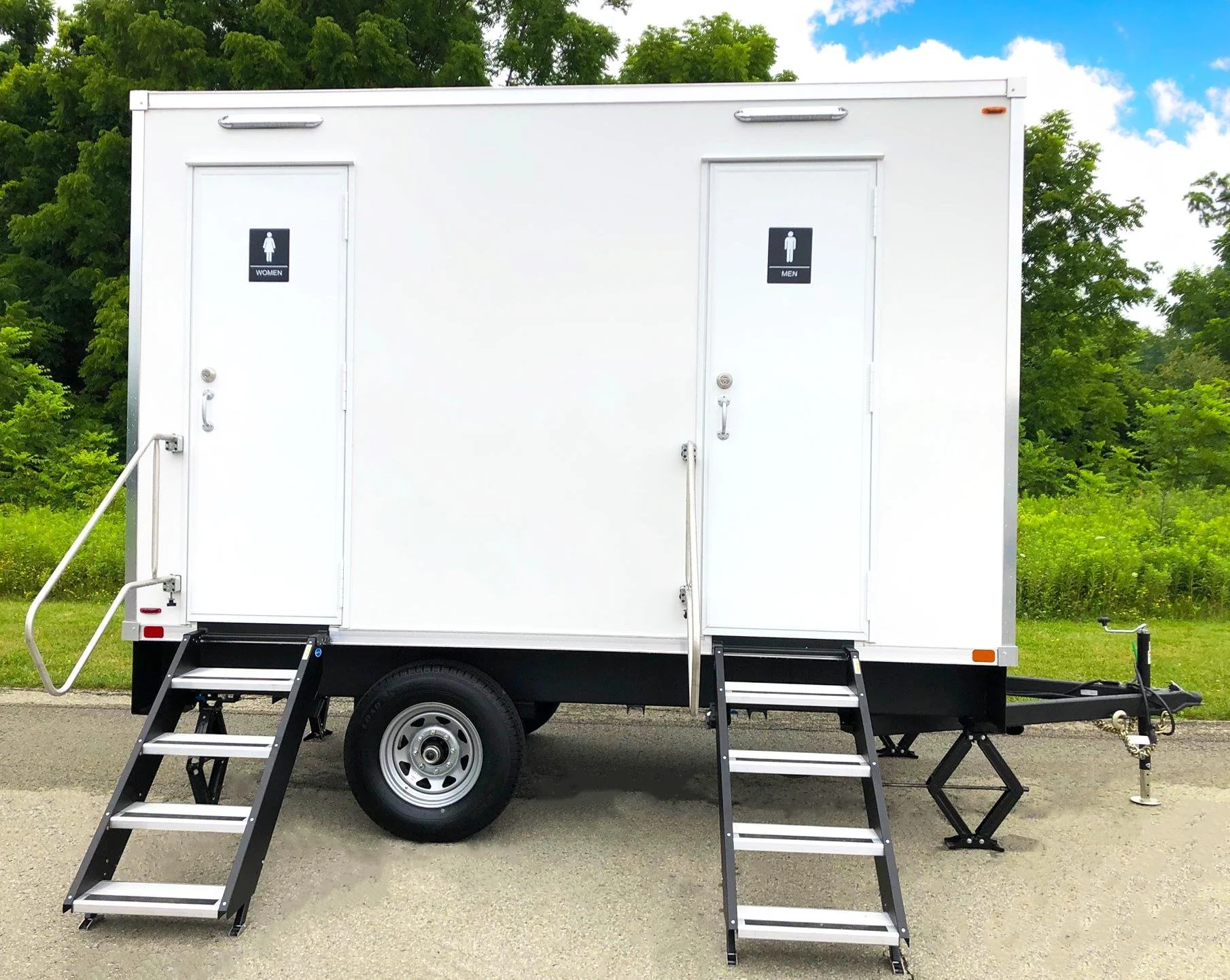 A mobile restroom trailer with separate entrances for women and men, each with a small set of stairs and a door, situated outdoors on a paved surface with green trees and a partly cloudy sky in the background.
