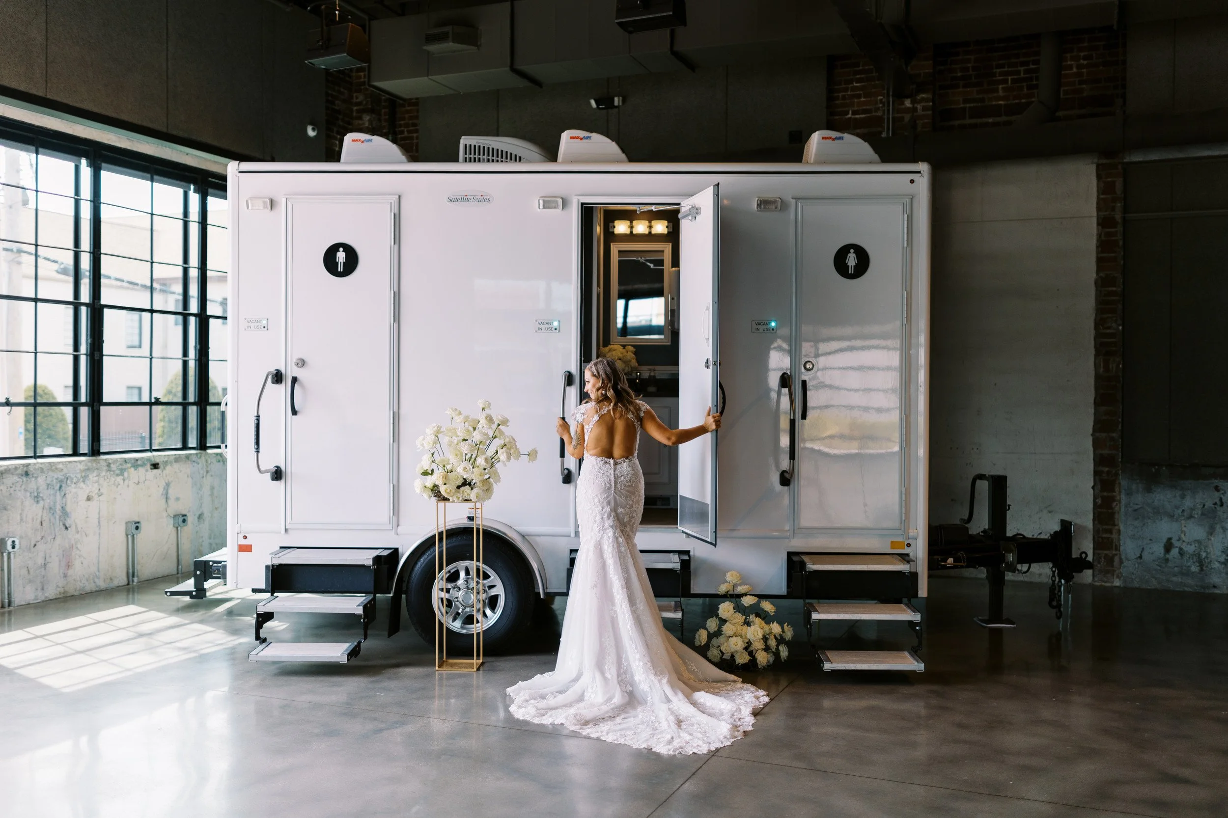 A bride in a wedding dress standing outside a portable restroom trailer, with steps and floral decorations nearby, inside a spacious industrial-style venue with large windows.