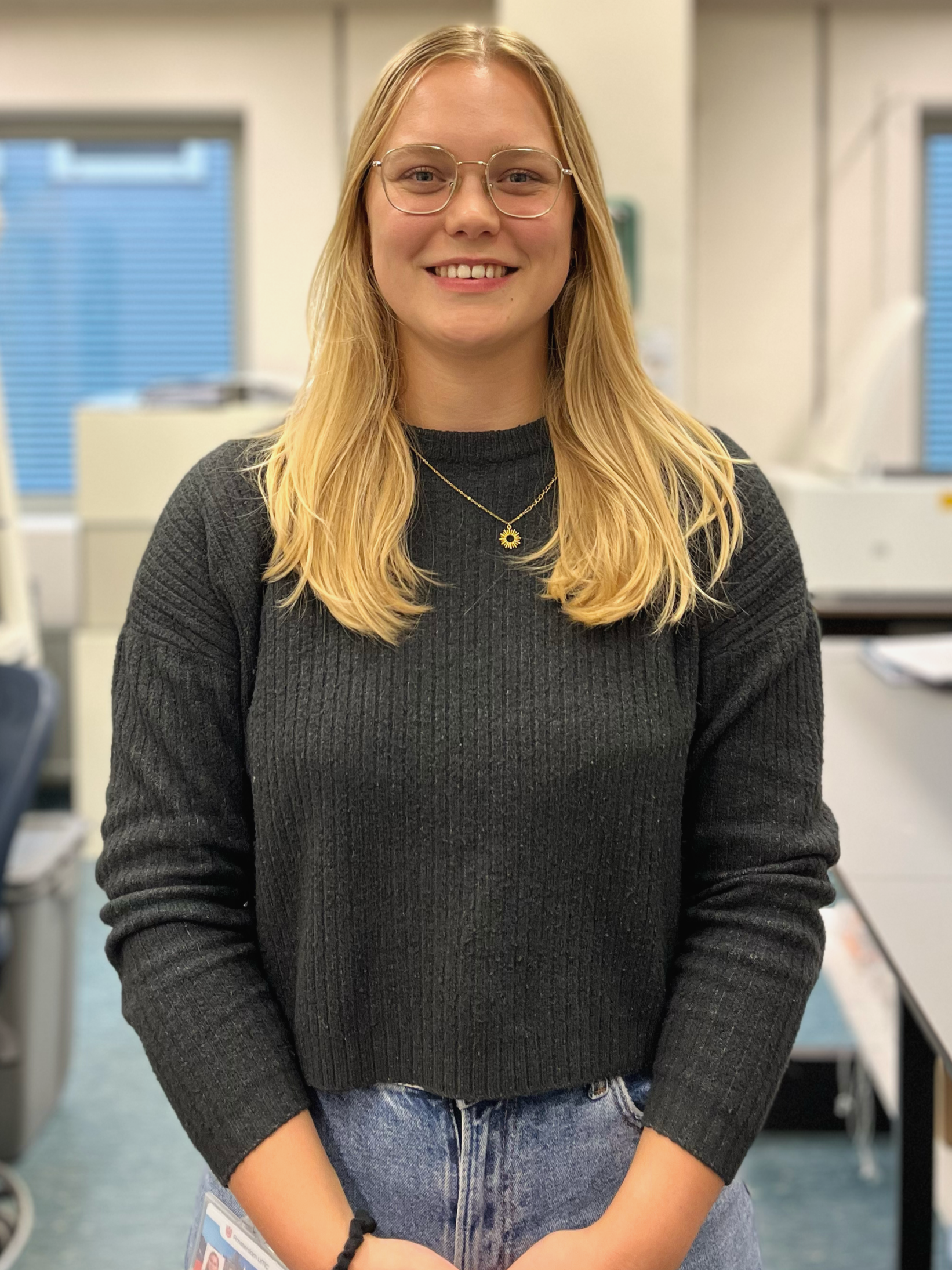 A young woman with long blonde hair, wearing glasses, a black sweater, and blue jeans, smiling in an indoor setting that appears to be an office.