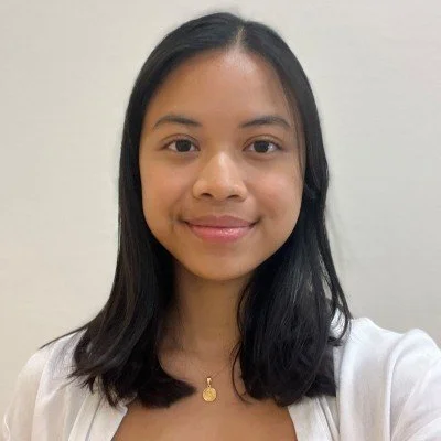 A young woman with straight, shoulder-length black hair, smiling, wearing a white top and a gold necklace with a circular pendant, standing against a plain white wall.