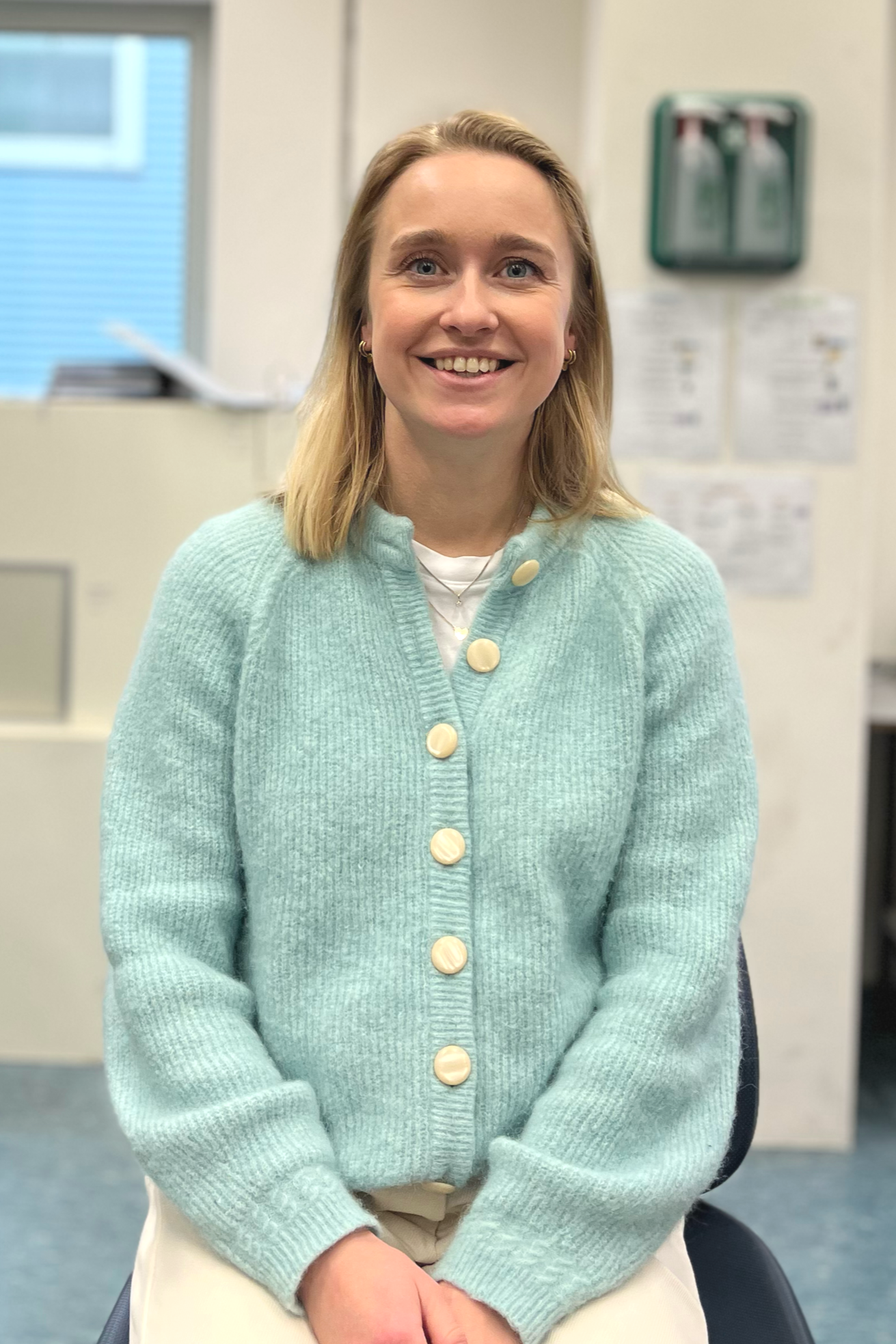 A woman sitting and smiling in an office, wearing a light blue cardigan with large buttons and beige pants. There are papers and a window in the background.