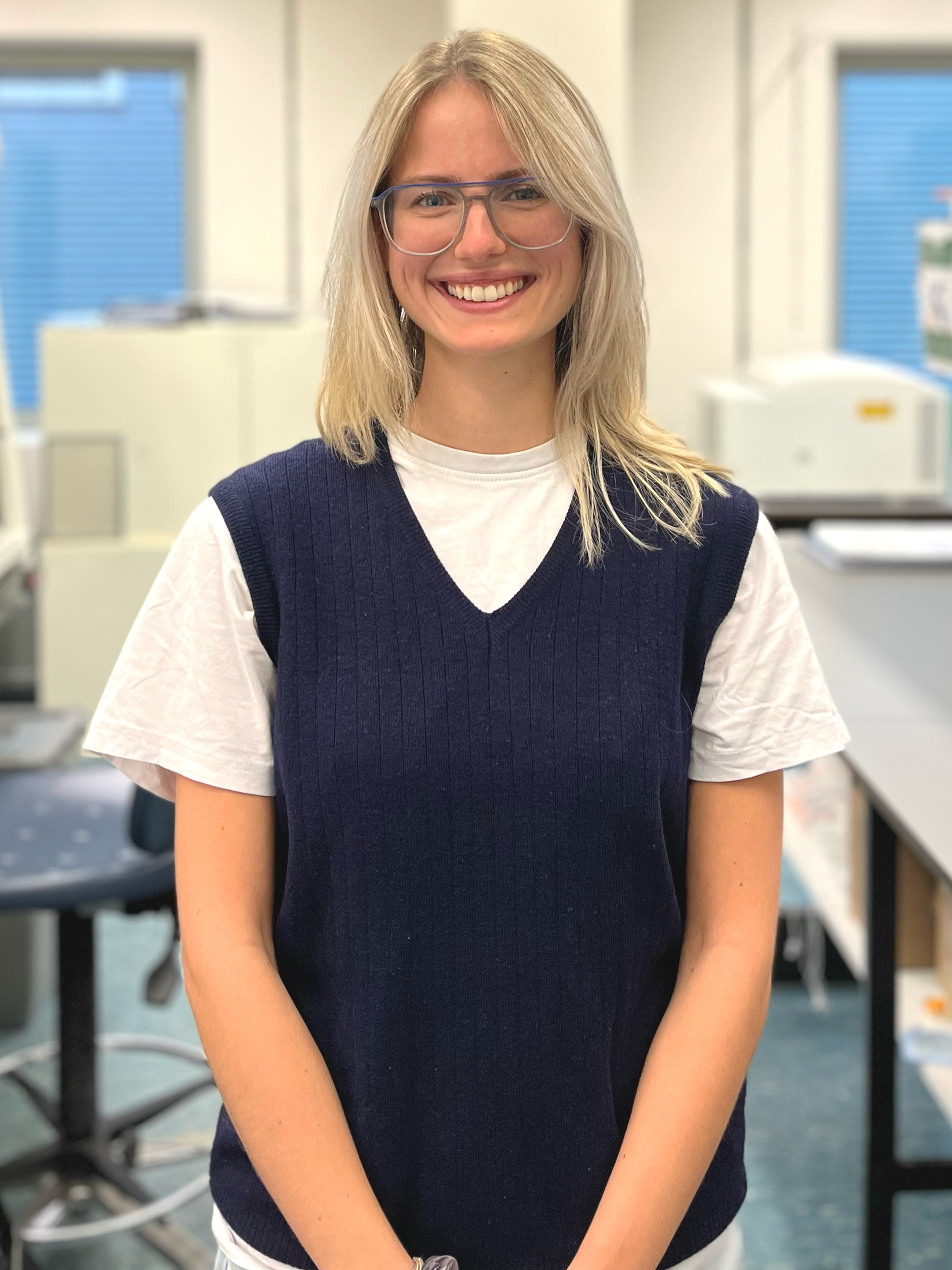 A young woman with shoulder-length blonde hair, wearing glasses, a white t-shirt, and a navy blue sweater vest, smiling in a bright office environment with desks and windows in the background.