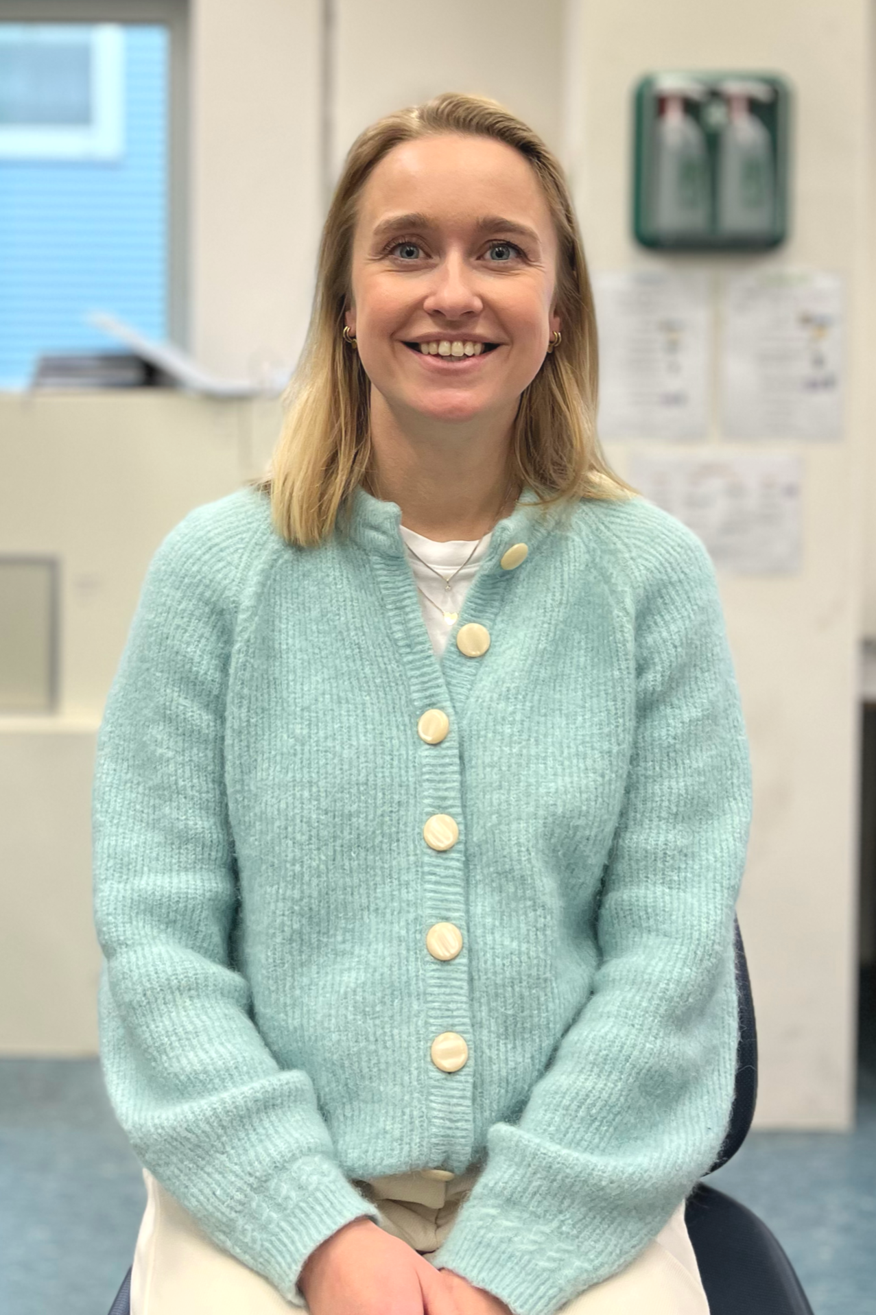 A young woman with blonde hair and blue eyes smiling, sitting in an office or classroom, wearing a light blue button-up cardigan and a white shirt.